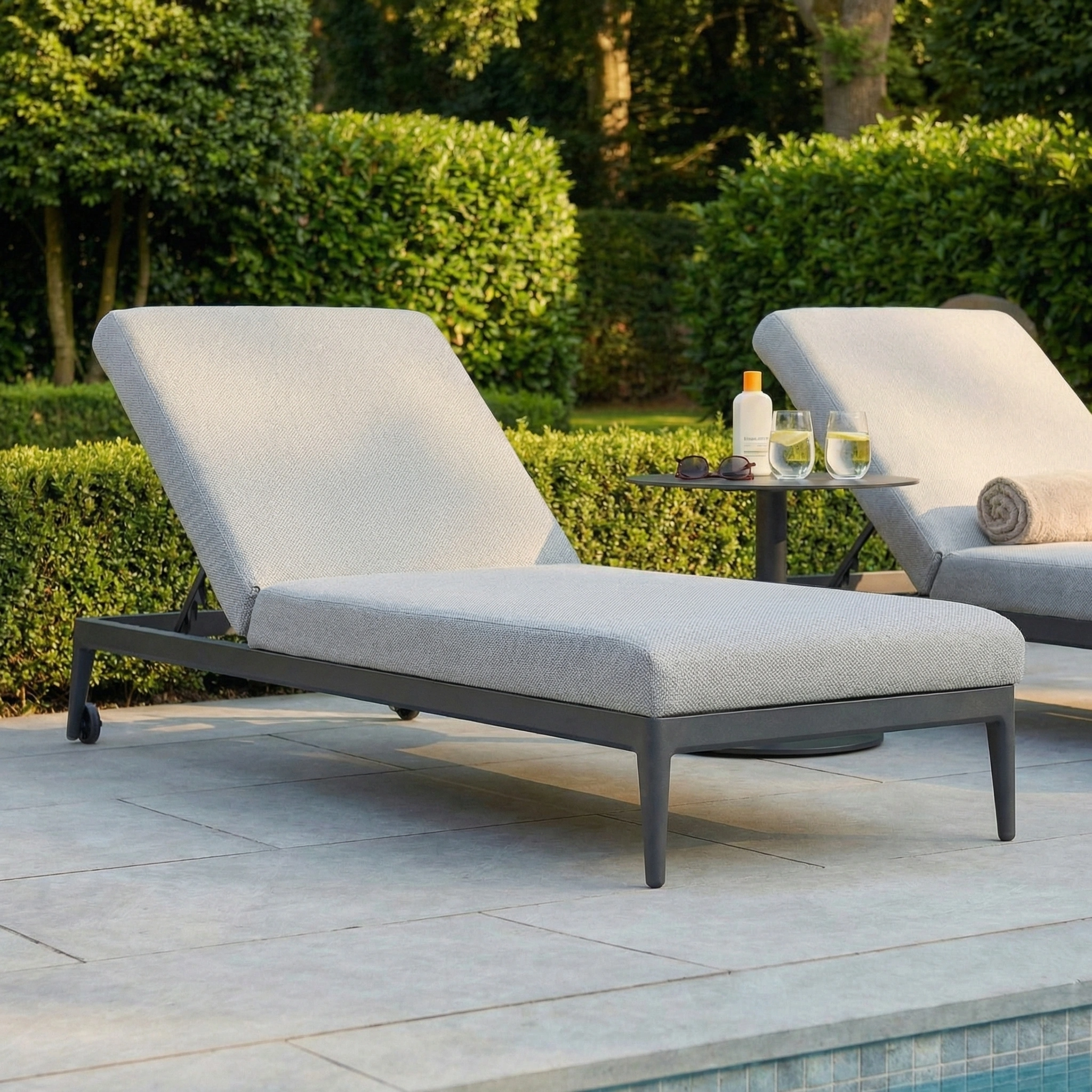 Luna Outdoor Fabric Sun Lounger in Oyster Grey by the pool is cushioned, with drinks, towels, and lush greenery in the background.