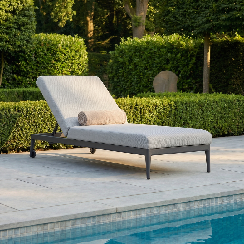 The Luna Outdoor Fabric Sun Lounger in Oyster Grey, topped with a beige rolled towel, is set on a stone patio by green hedges.