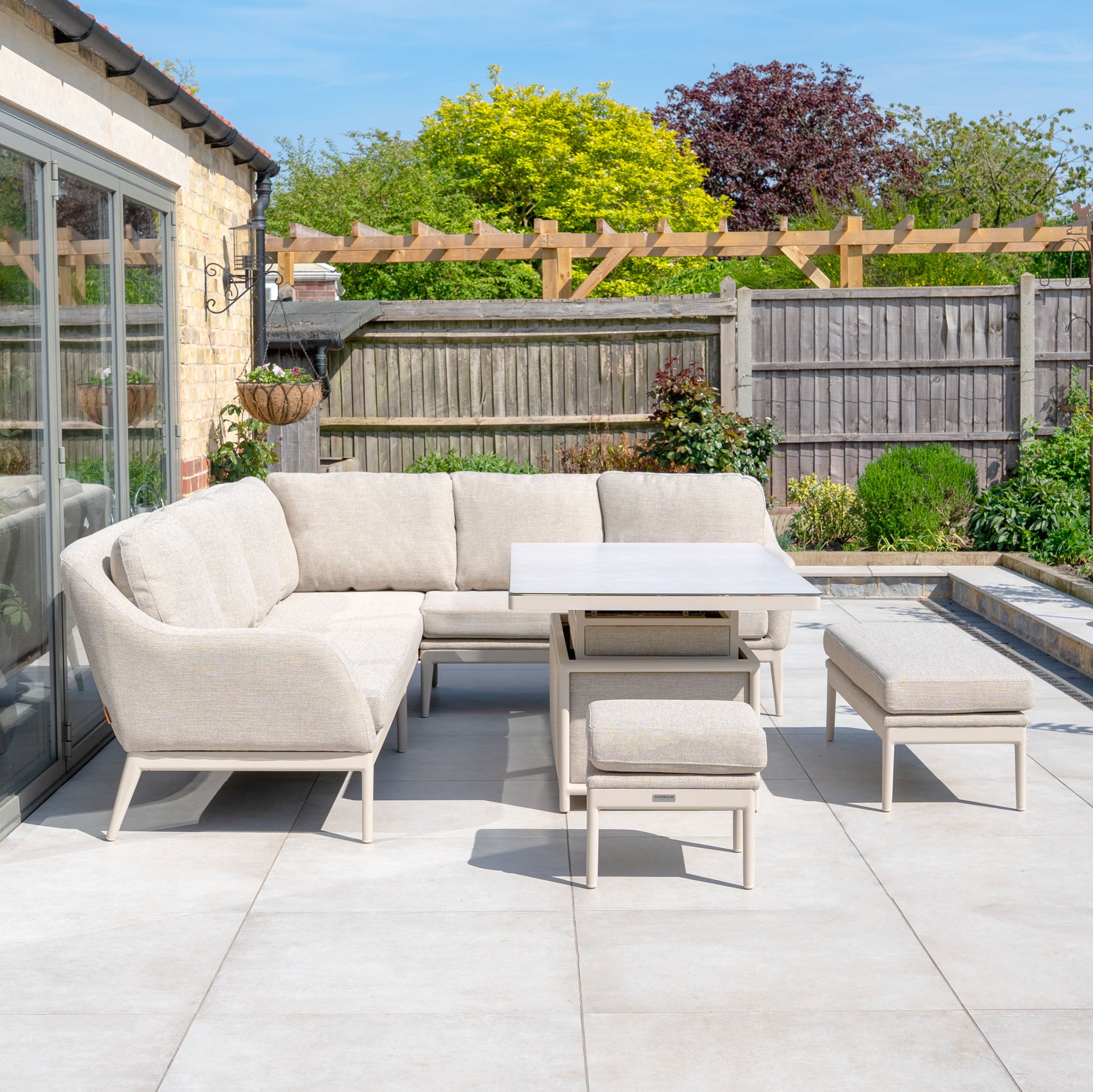 The Luna Outdoor Fabric Rectangular Corner Dining Set with Rising Table in Fawn (Left Hand) is shown on a tiled patio, featuring durable fabric, with garden plants and a wooden fence in the background.
