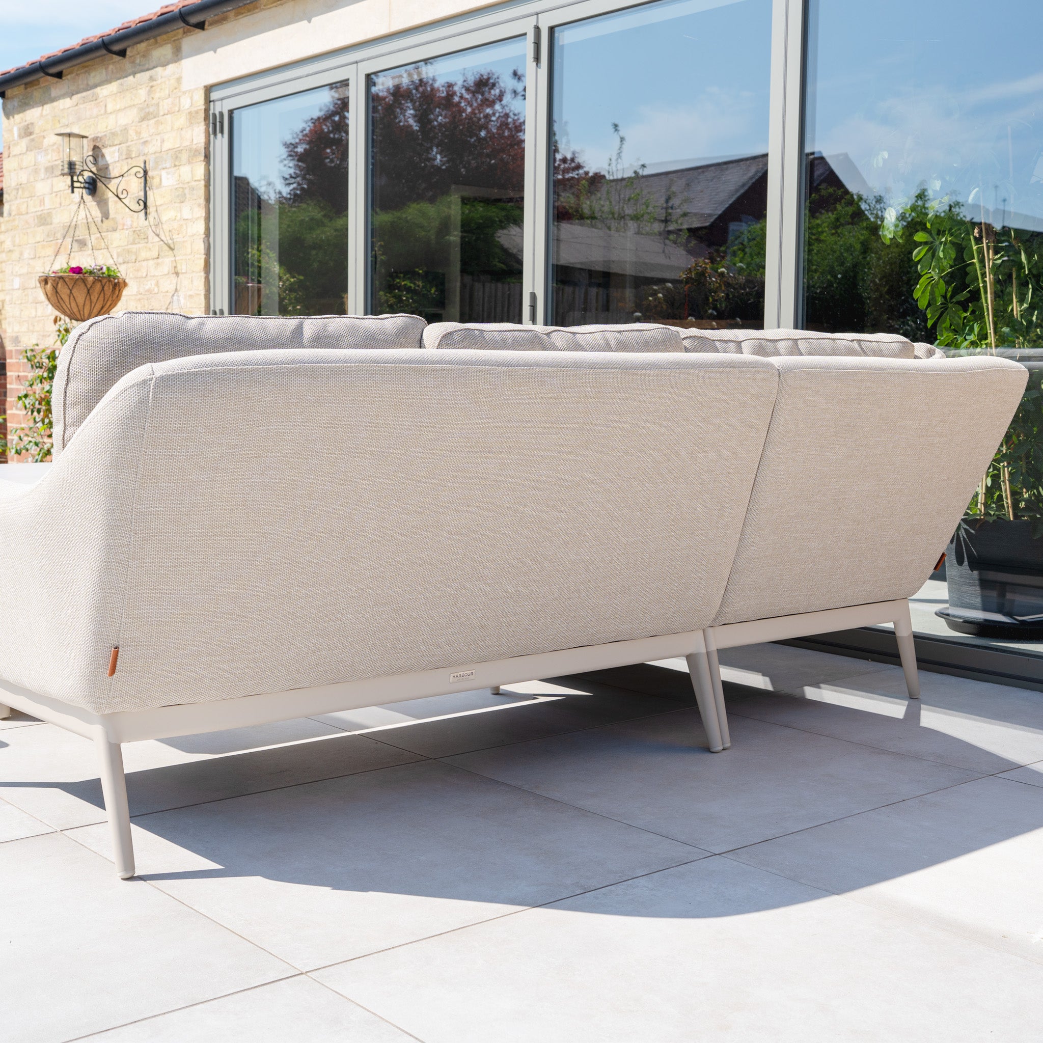The Luna Outdoor Fabric Rectangular Corner Dining Set with Rising Table in Fawn (Left Hand) is shown on a tiled patio beside large glass doors, surrounded by plants and a sunlit stone wall.