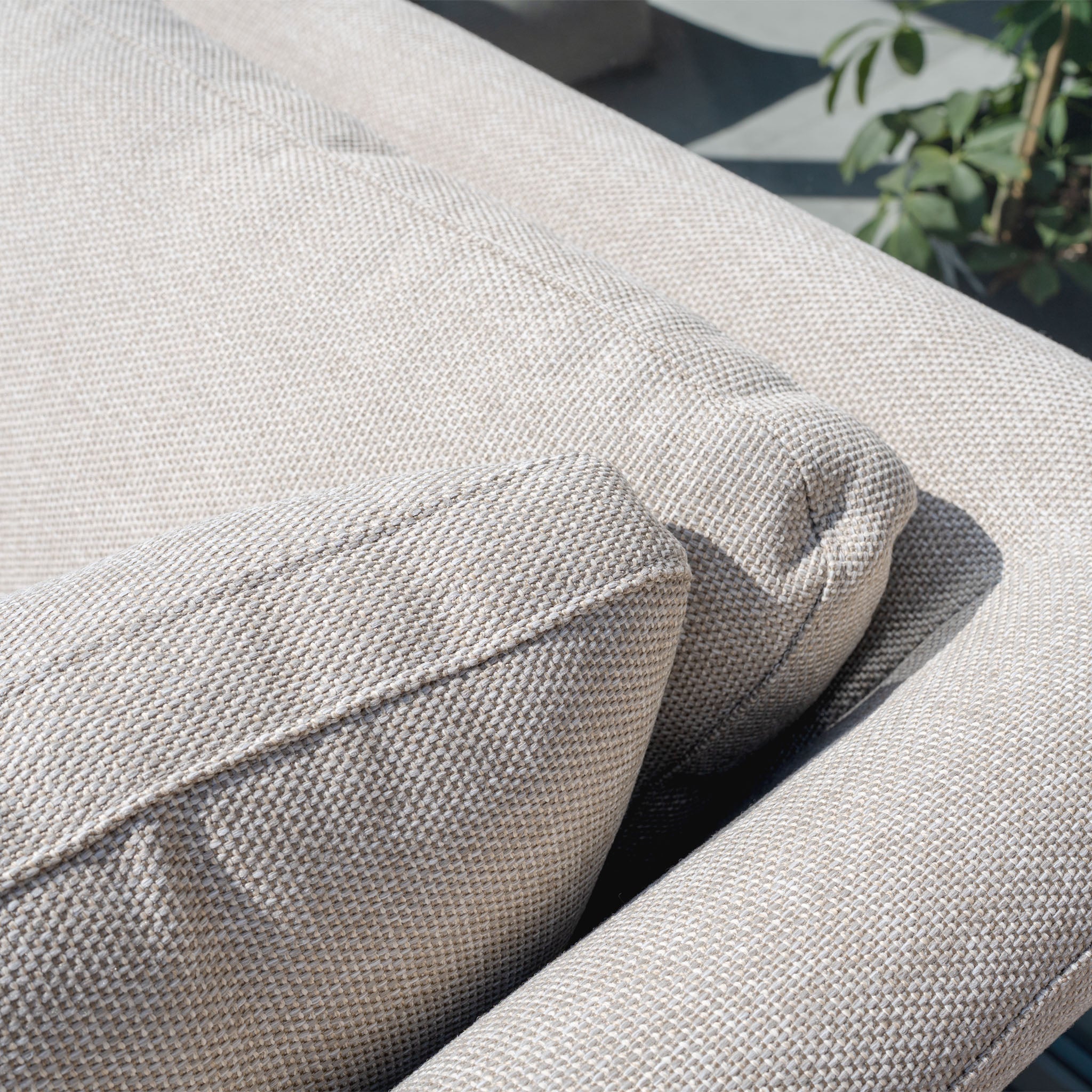 Close-up of the Luna Outdoor Fabric Rectangular Corner Dining Set with Rising Table in Fawn (Left Hand), featuring light beige textured fabric, sunlight, and a leafy plant in the background.