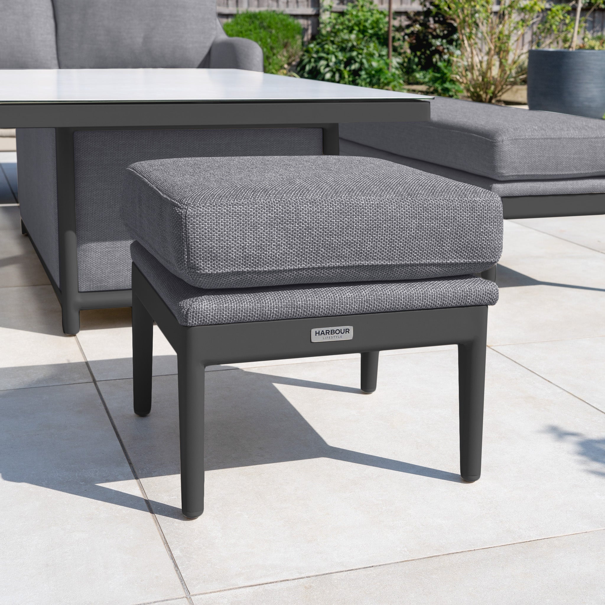 The Luna Stool in Grey, with cushion, sits on a tiled patio beside outdoor furniture and a table, with plants in the background. This stylish all-weather stool is perfect for relaxing outdoors.