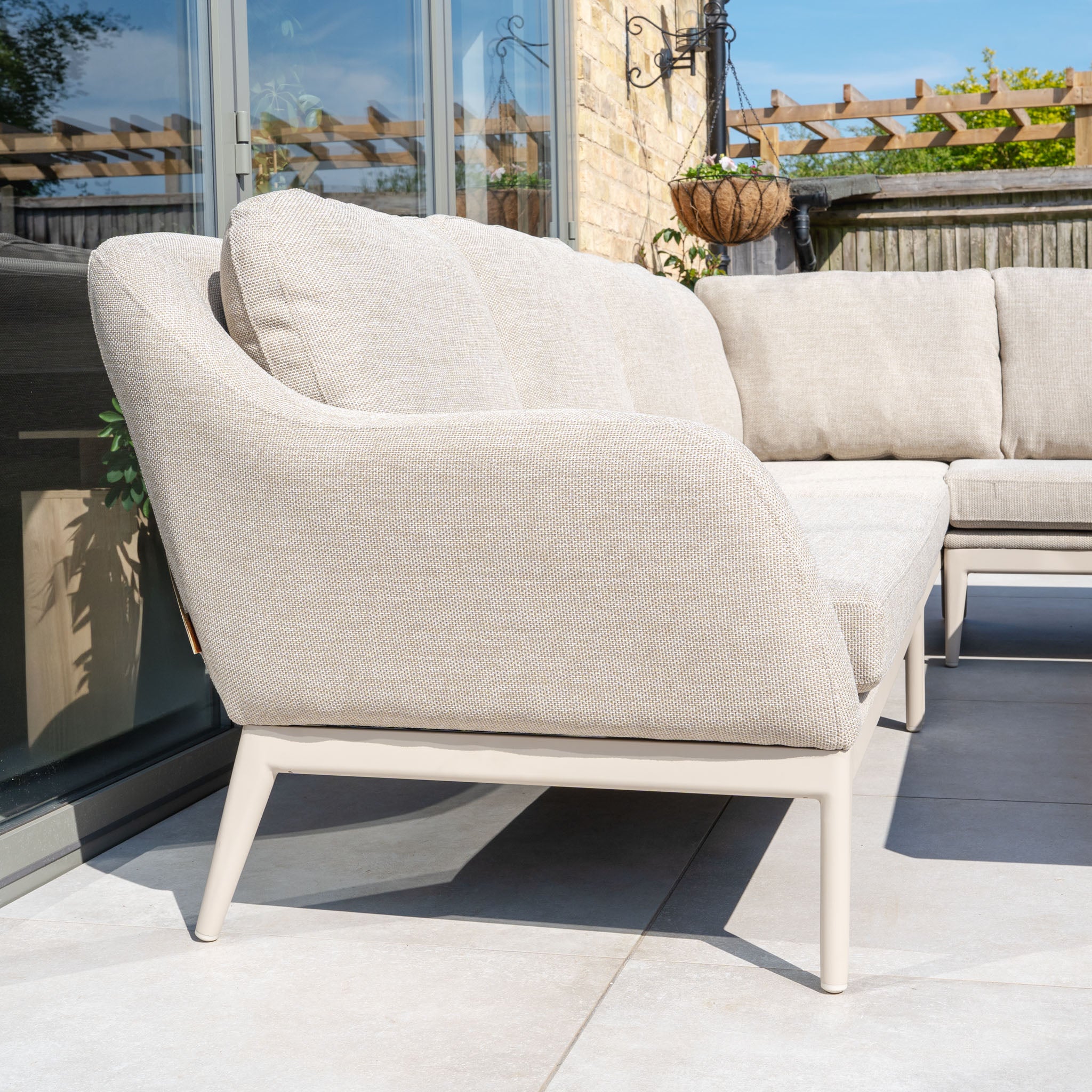 The Luna Outdoor Fabric Rectangular Corner Dining Set with Rising Table in Fawn (Left Hand) features light beige fabric and maintenance-free metal legs, perfect for a patio by glass doors and a brick wall.