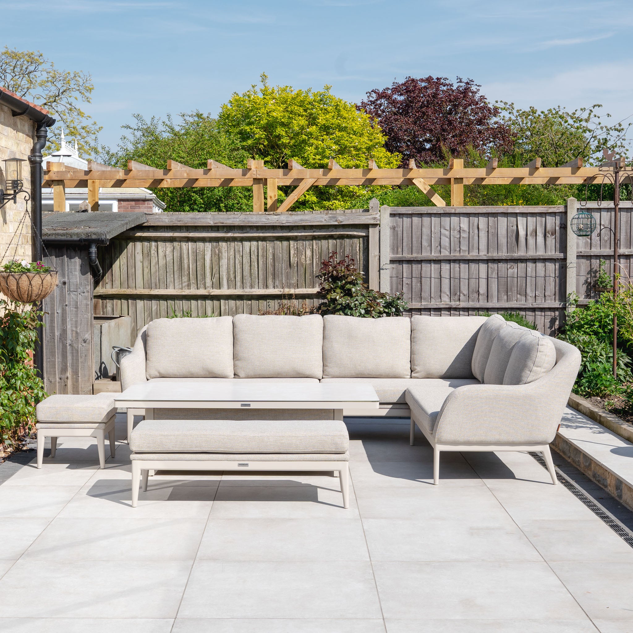 The Luna Outdoor Fabric Rectangular Corner Dining Set with Rising Table in Fawn (Right Hand) sits on a tiled patio, complemented by a garden fence and lush greenery in the background.