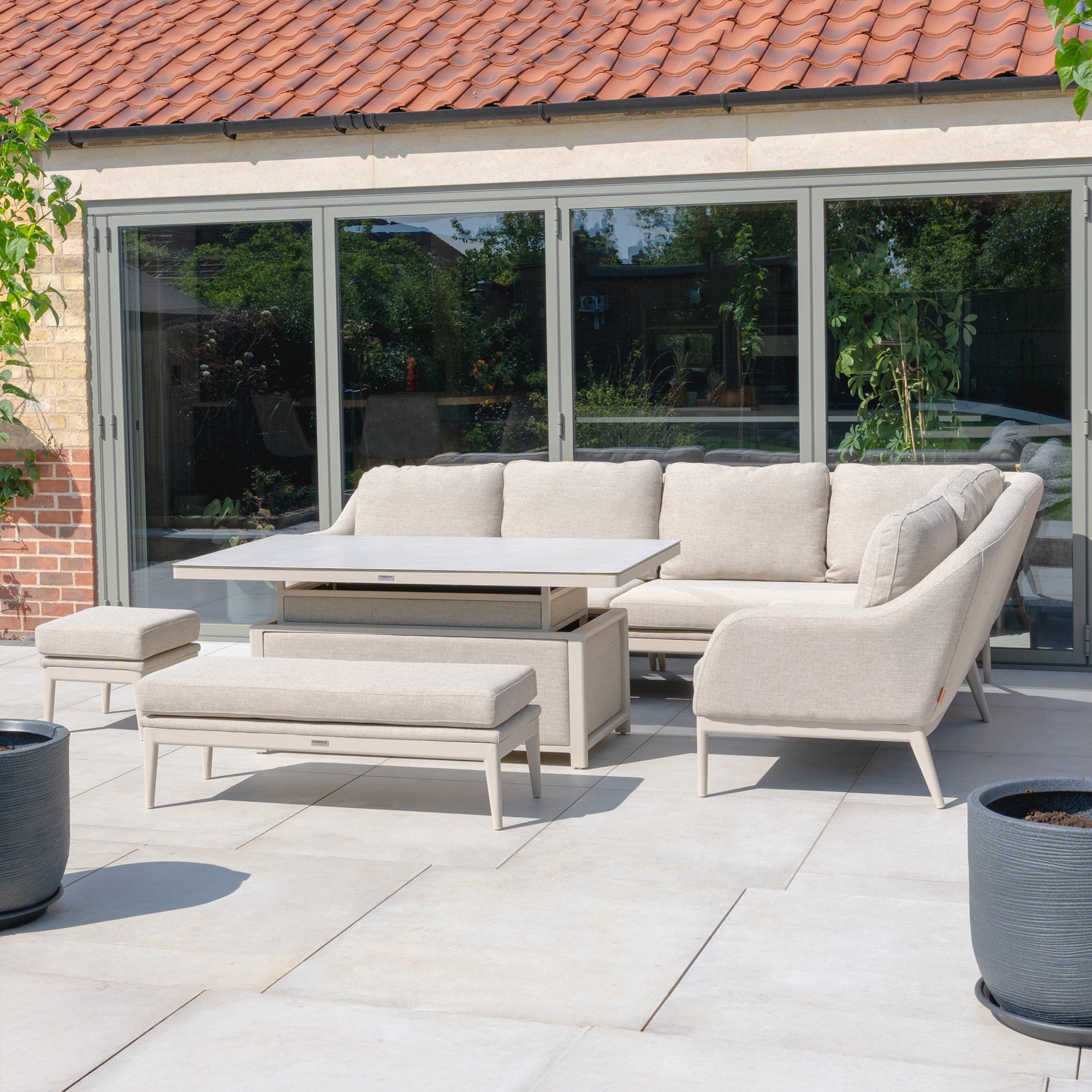 The Luna Outdoor Fabric Rectangular Corner Dining Set with Rising Table in Fawn (Right Hand) features beige cushions, a glass rising table, and a sleek aluminium frame, perfect for patios by glass doors.