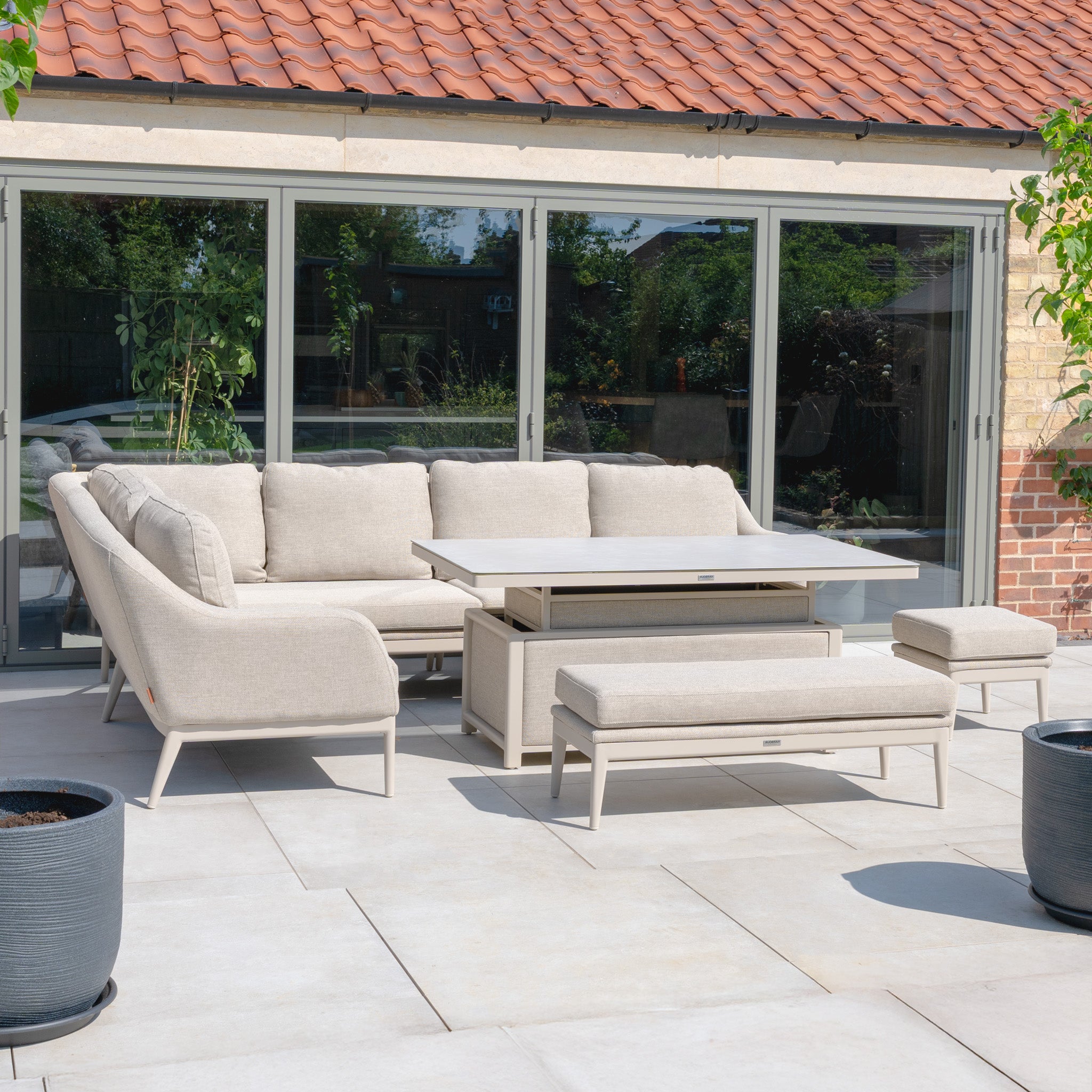 The Luna Outdoor Fabric Rectangular Corner Dining Set with Rising Table in Fawn (Left Hand) features a beige sectional, benches, and a rising table—perfect for a modern patio by the glass doors.