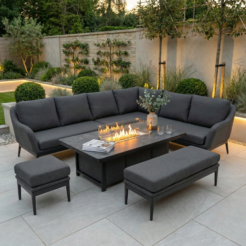 The Luna Outdoor Fabric Rectangular Corner Dining Set with Rising Firepit Table in Grey (Right Hand) creates a cozy patio spot with gray cushions and a rising firepit table, surrounded by lush plants in the background.