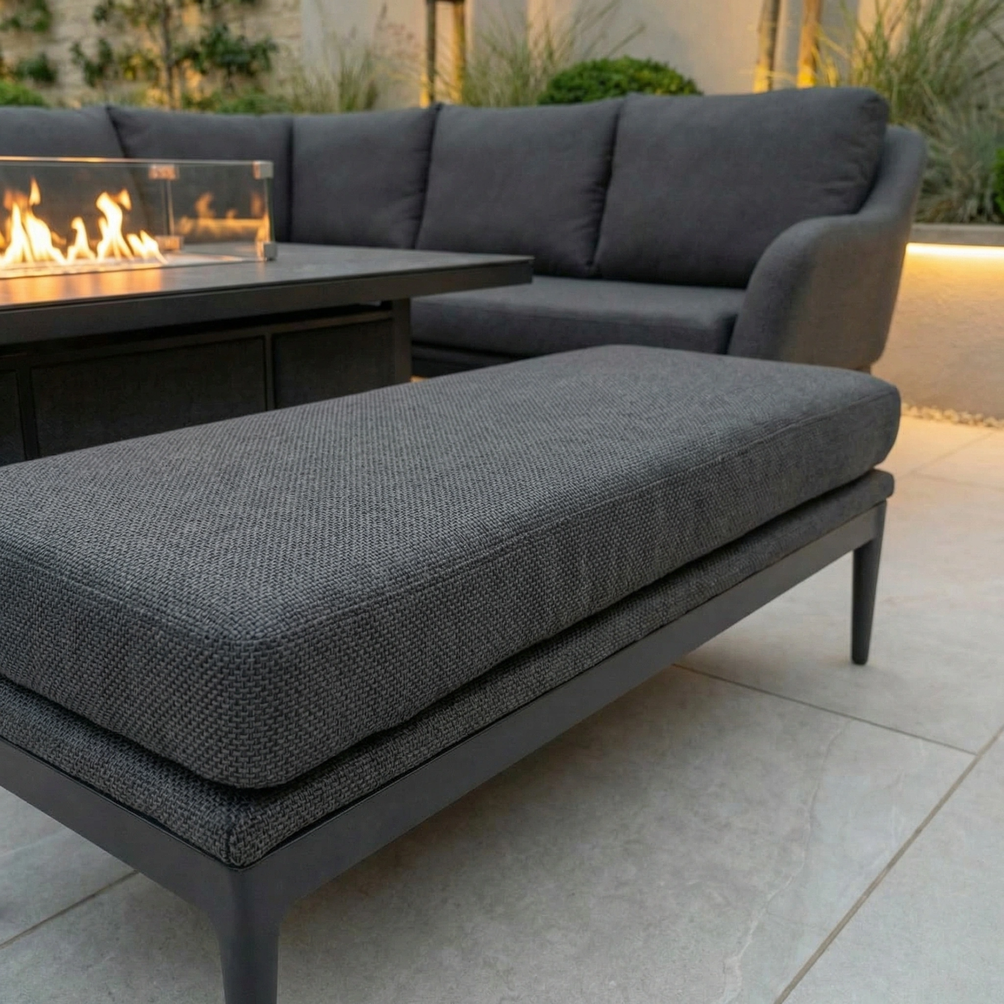 The Luna Outdoor Fabric Rectangular Corner Dining Set with Rising Firepit Table in Grey (Right Hand) sits on a tiled patio surrounded by lush plants.
