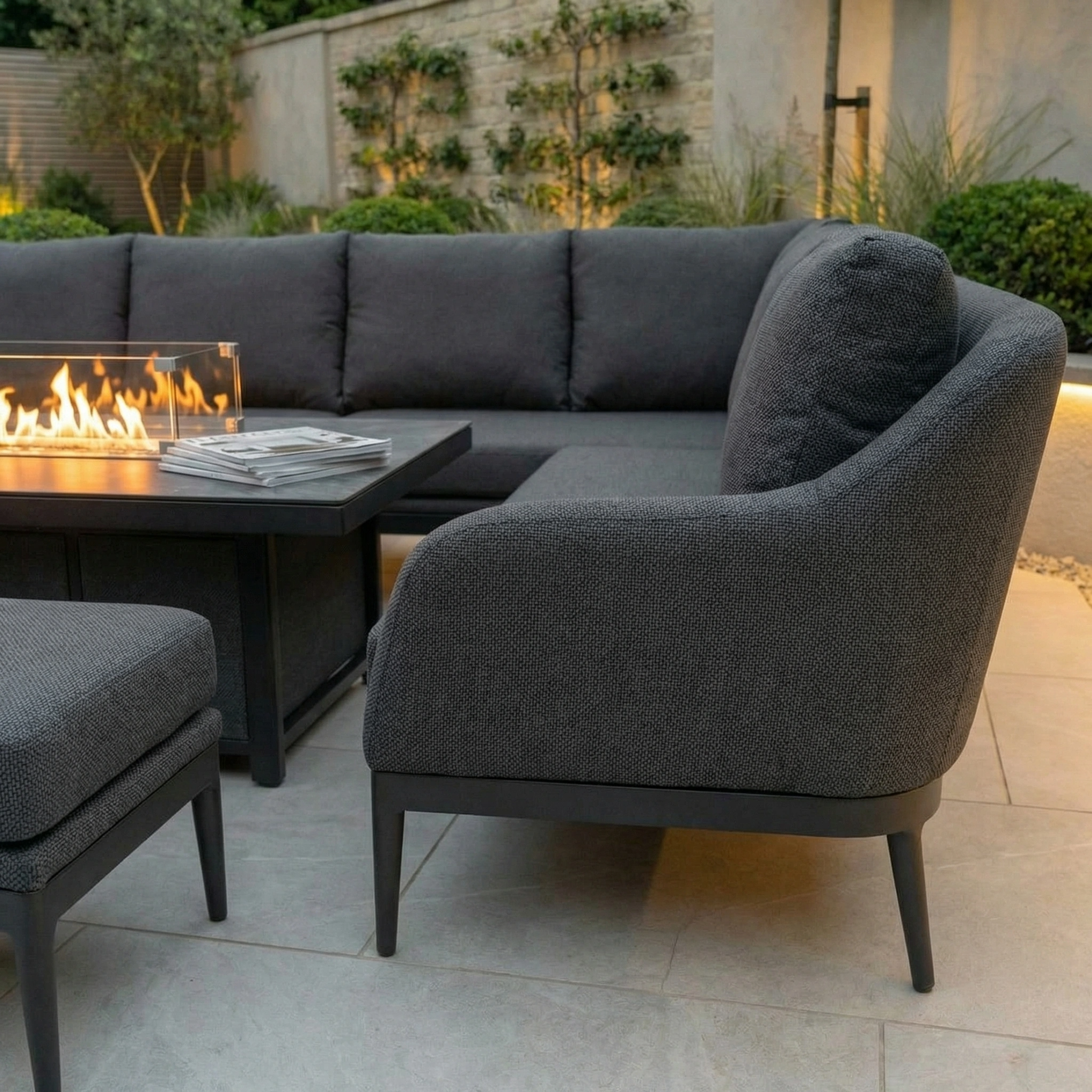 Modern outdoor patio featuring the Luna Outdoor Fabric Rectangular Corner Dining Set with Rising Firepit Table in Grey (Right Hand), stylish seating, and lush greenery in the background.