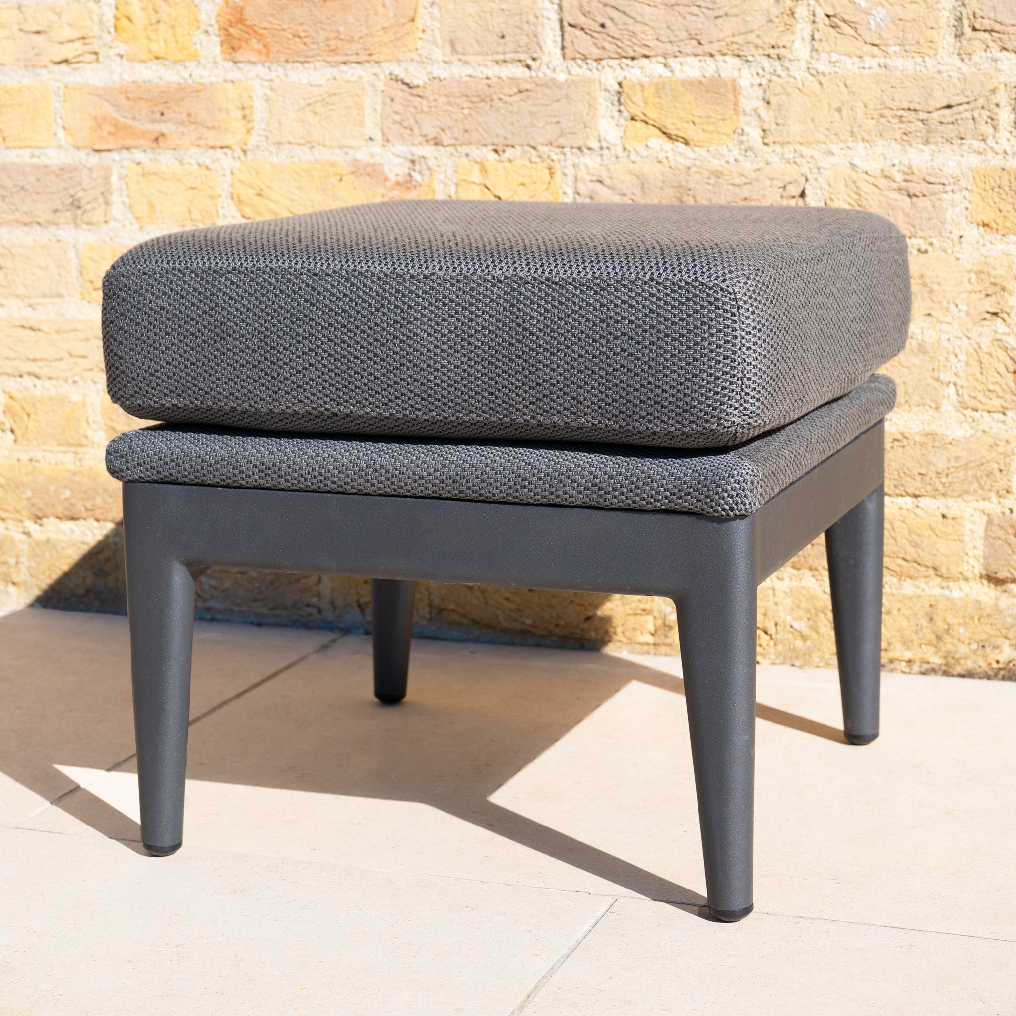 The Luna Stool in Grey, featuring sleek black legs, is set on a patio against a yellow brick wall, making it an ideal choice for stylish outdoor seating.