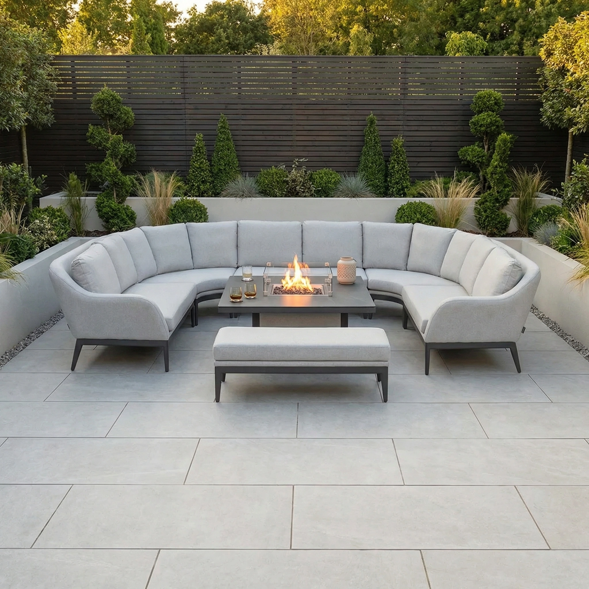 The Luna U-Shape Outdoor Fabric Corner Dining with Rising Firepit Table in Oyster Grey pairs with lush greenery and a dark wooden fence to create a stylish and inviting modern patio space.