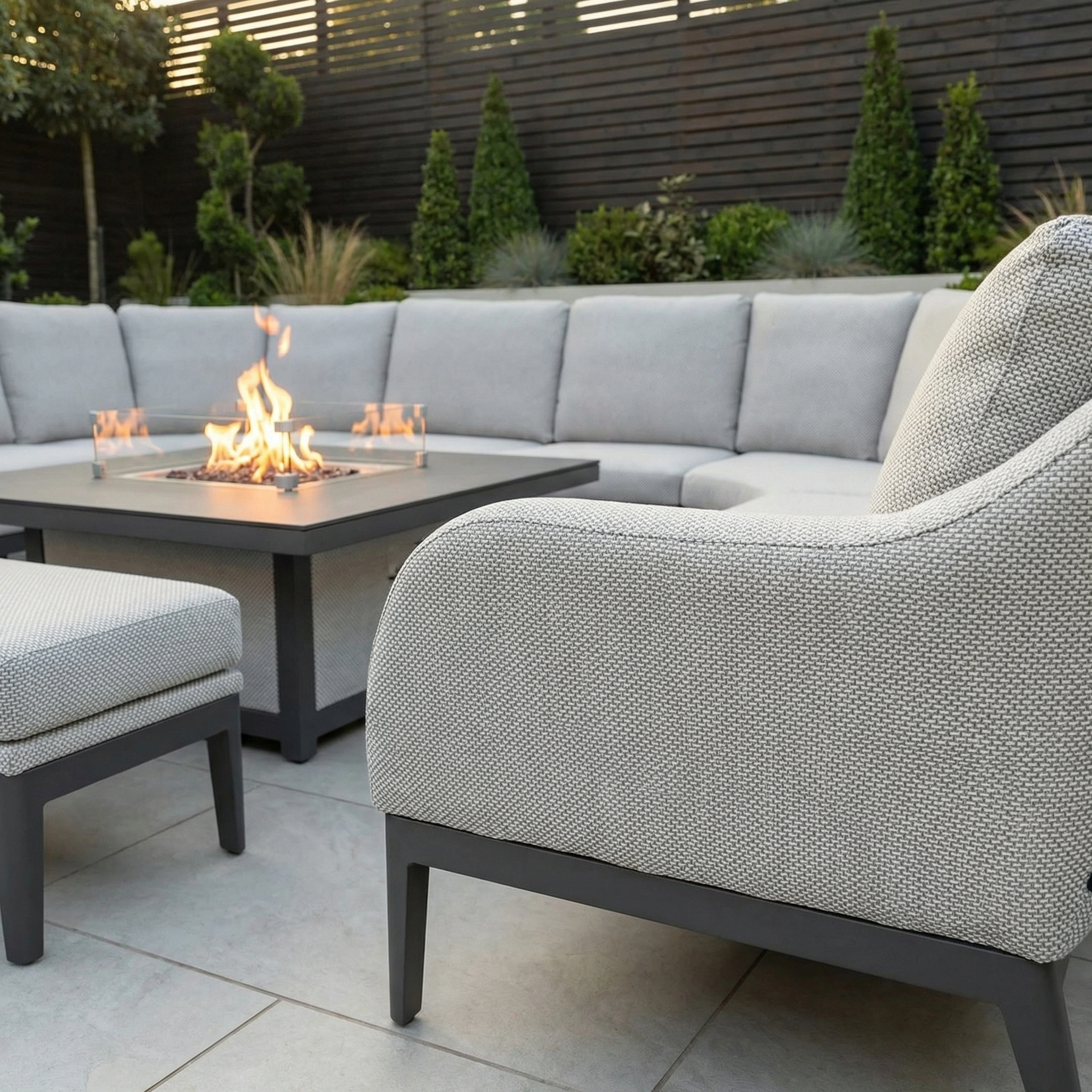 Contemporary patio with Luna U-Shape Outdoor Fabric Corner Dining in Oyster Grey, a rising firepit table, and vibrant green landscaping in the background.