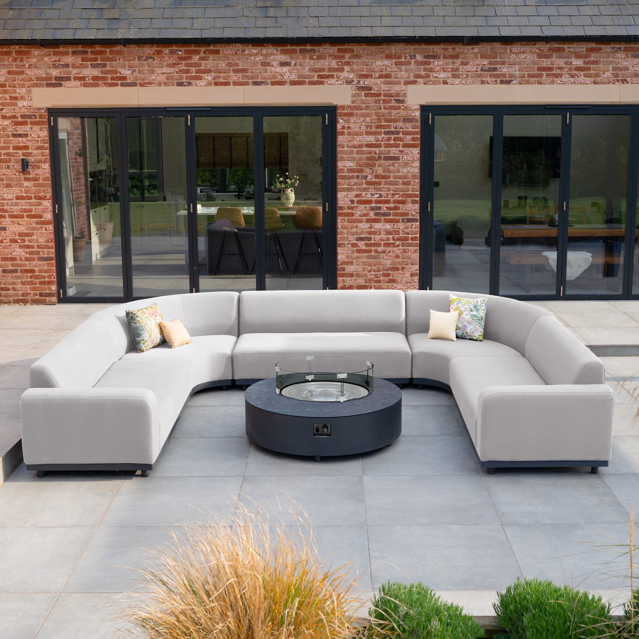 The Luna U-Shape Outdoor Fabric Curved Corner Group with Round Firepit Coffee Table in Oyster Grey creates a stylish outdoor living space on a modern patio in front of a brick house.