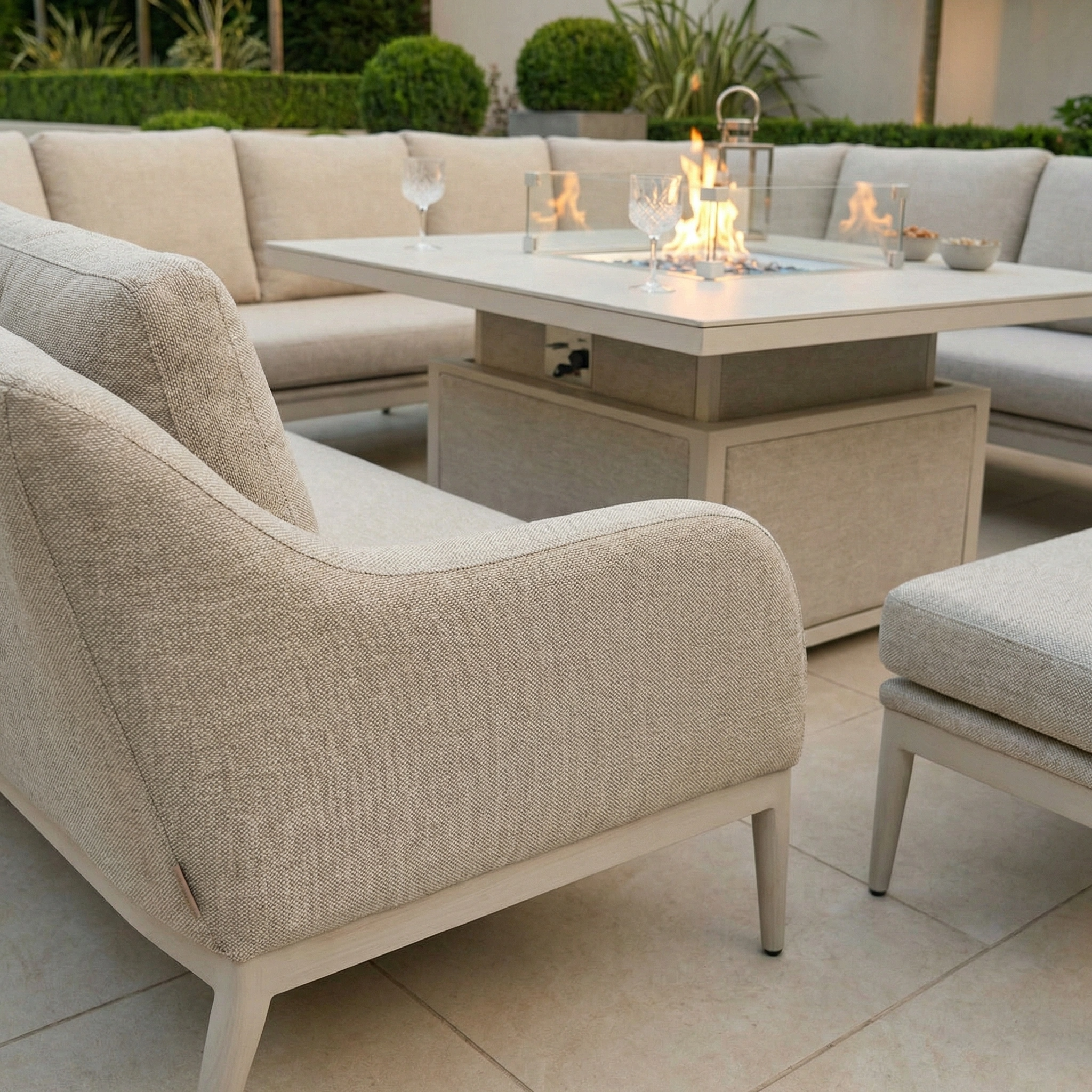 The Luna U-Shape Outdoor Fabric Corner Dining with Rising Table in Fawn set features a beige sofa arranged around a height-adjustable table, with glasses and plants displayed in the background.