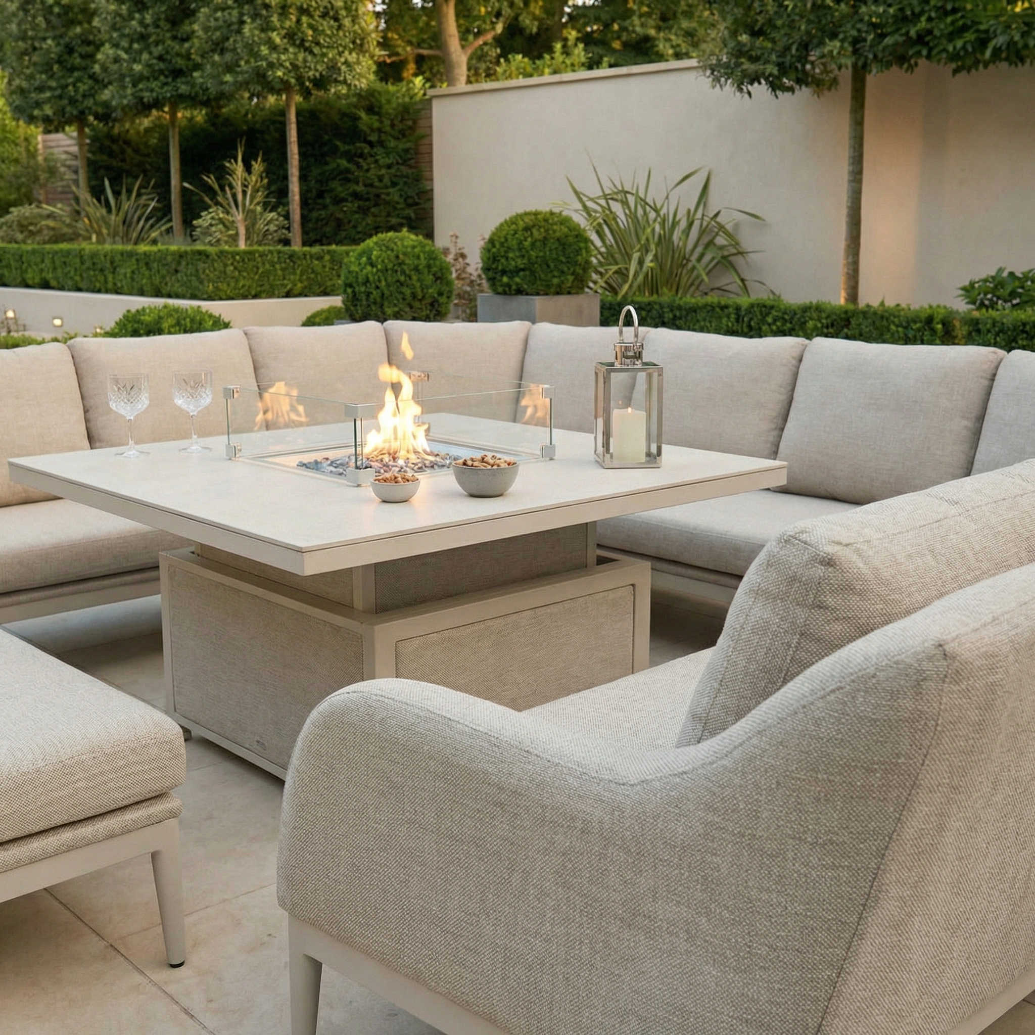 Modern patio featuring the Luna U-Shape Outdoor Fabric Corner Dining set in Fawn with a rising table, gray cushions, snacks, drinks, and lush greenery.
