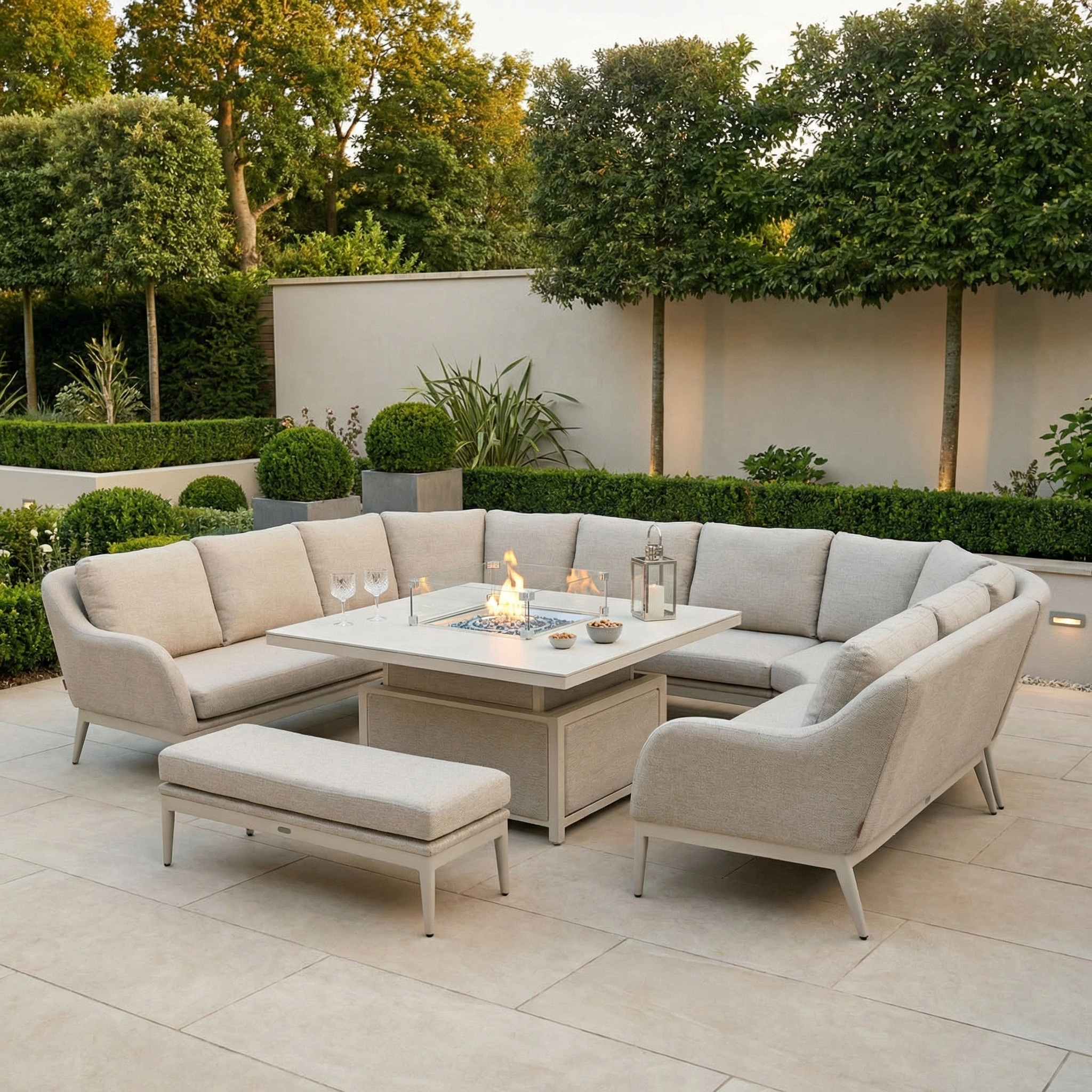 The Luna U-Shape Outdoor Fabric Corner Dining with Rising Table in Fawn features light grey cushions arranged around a height-adjustable firepit table, creating a stylish patio set amidst lush greenery.