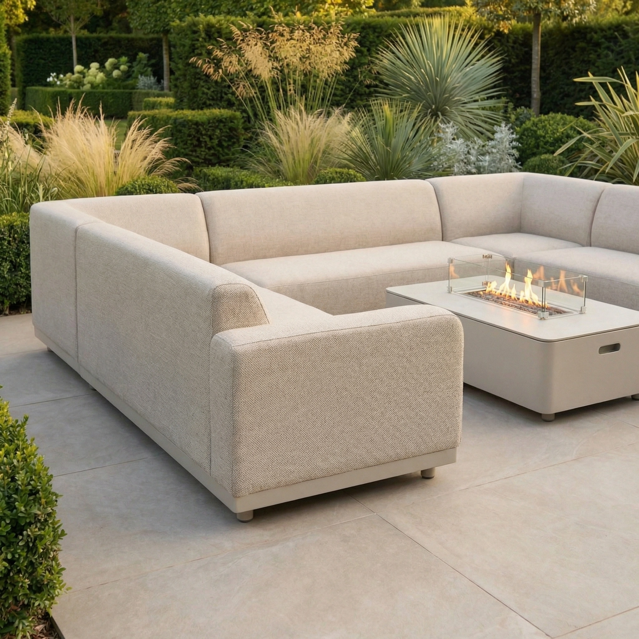 The Luna U-Shape Outdoor Fabric Sofa Set with Firepit Coffee Table in Fawn sits on a tiled patio, surrounded by lush green plants.