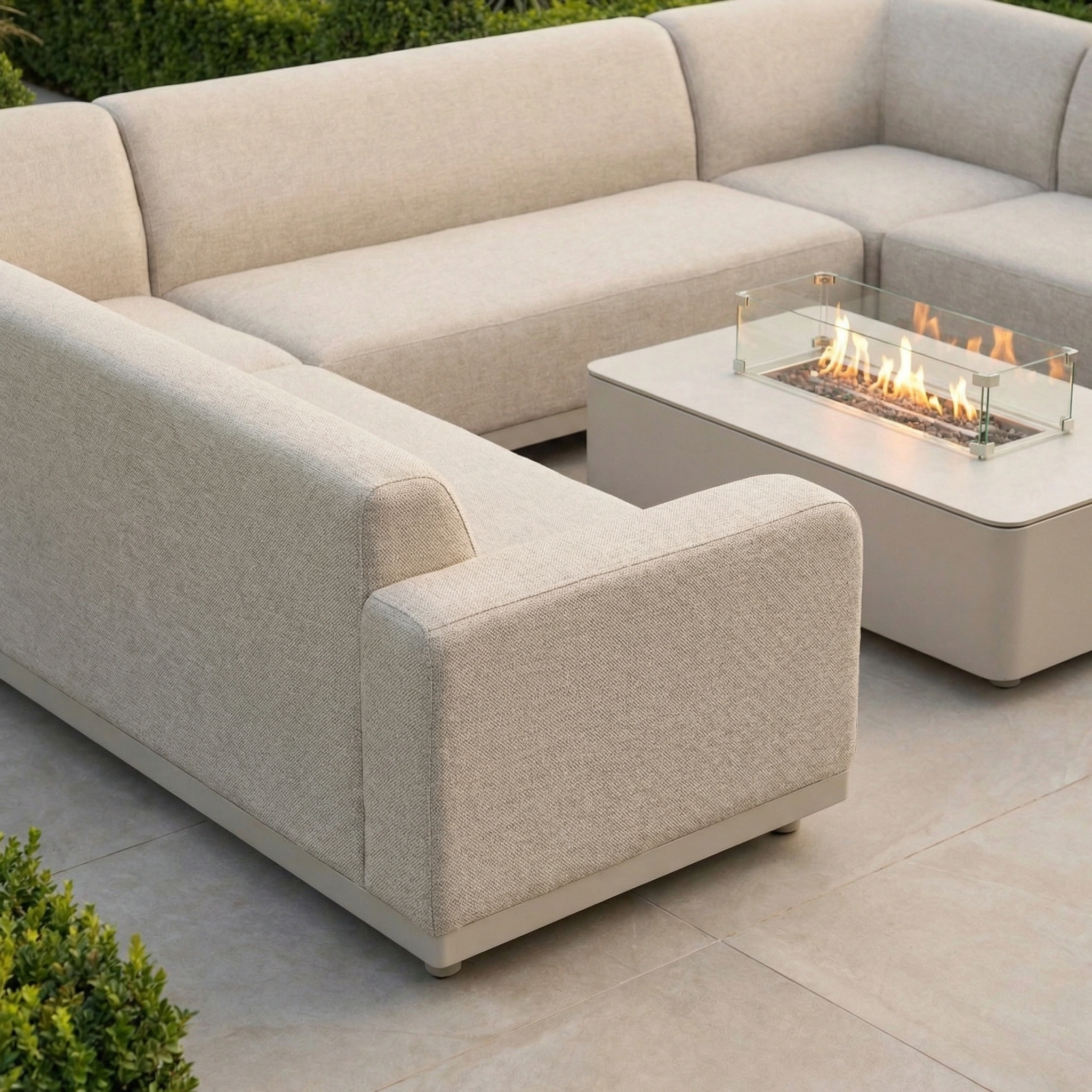The Luna U-Shape Outdoor Fabric Sofa Set with Firepit Coffee Table in Fawn offers weatherproof comfort and warmth, perfect for relaxing outdoors surrounded by greenery.