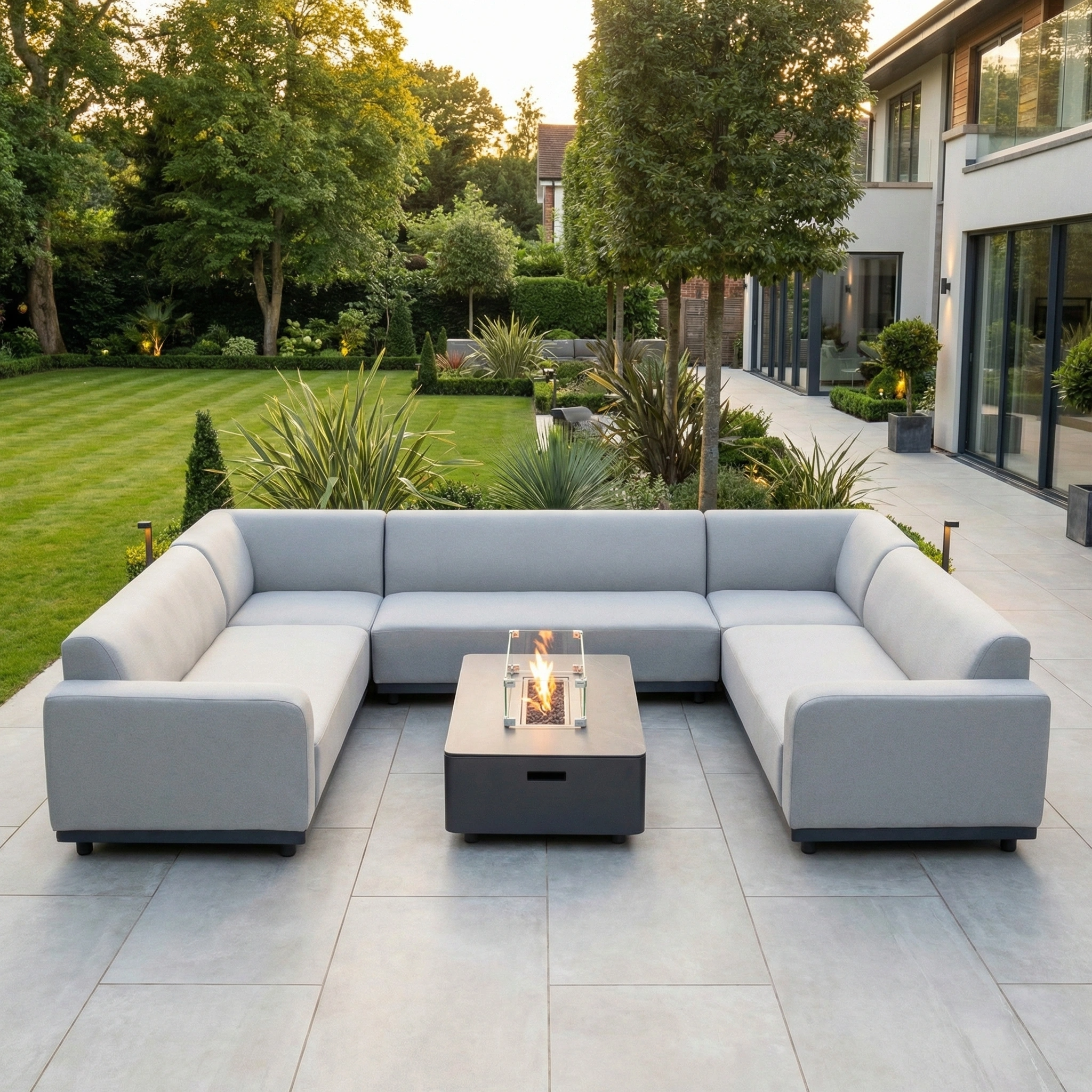 A modern patio features the Luna U-Shape Outdoor Fabric Sofa Set with Firepit Coffee Table in Oyster Grey, set amid greenery and a lush lawn.