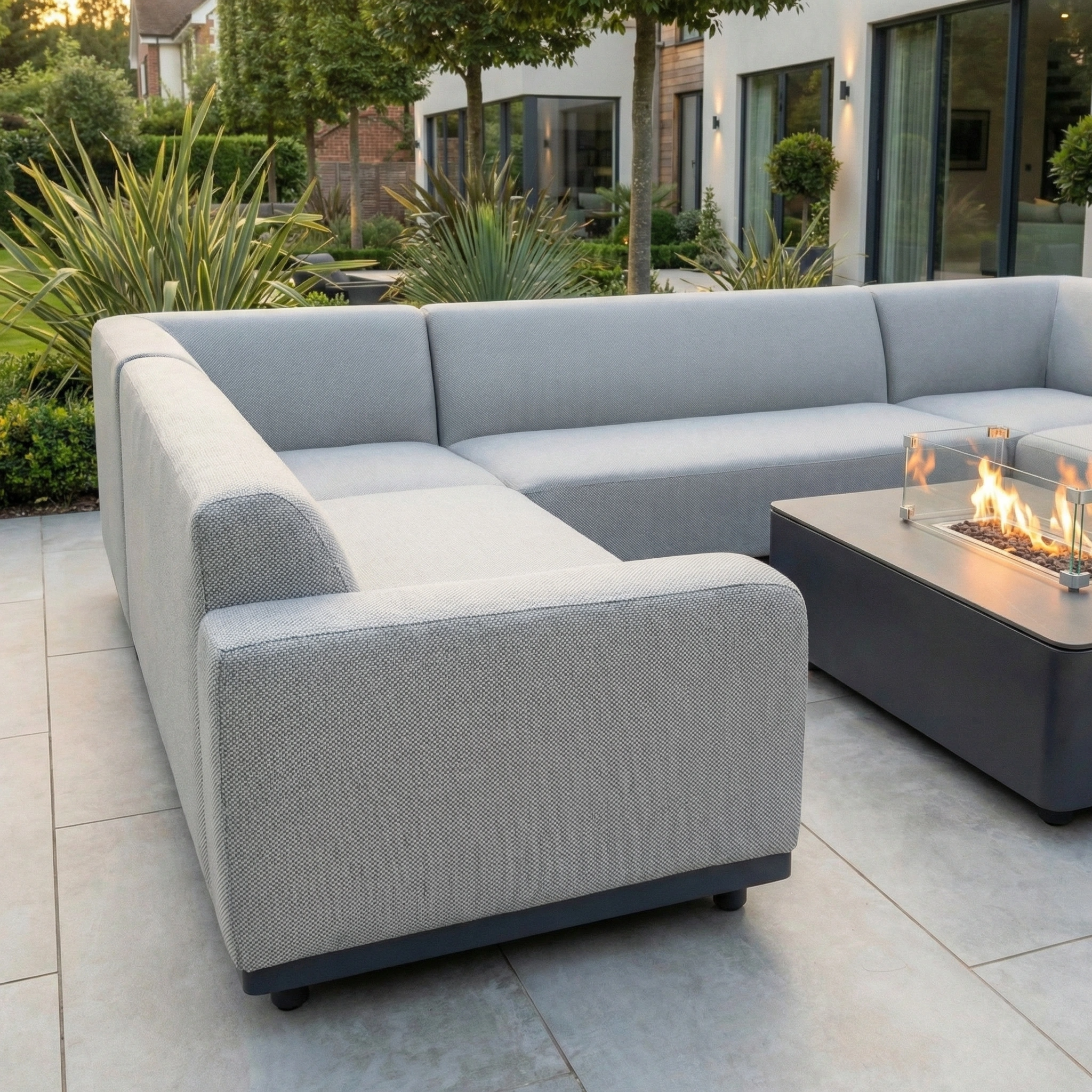 The Luna U-Shape Outdoor Fabric Sofa Set with Firepit Coffee Table in Oyster Grey creates a stylish, cozy retreat, accented by lush green plants.