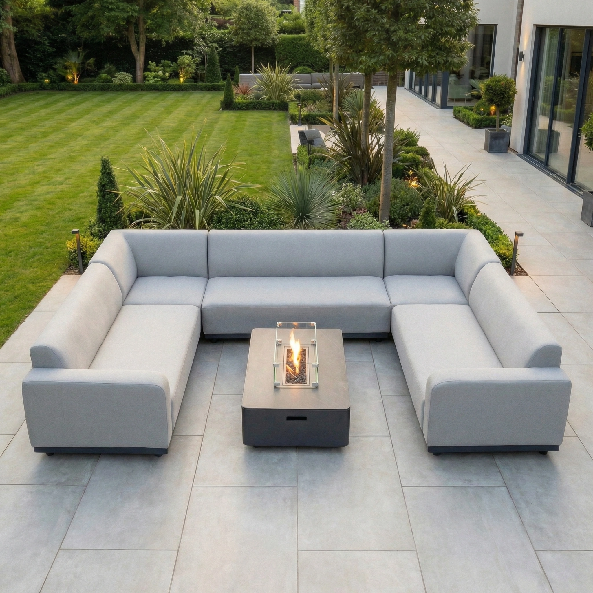 Modern outdoor patio featuring the Luna U-Shape Outdoor Fabric Sofa Set with Firepit Coffee Table in Oyster Grey, surrounded by a lush green garden.