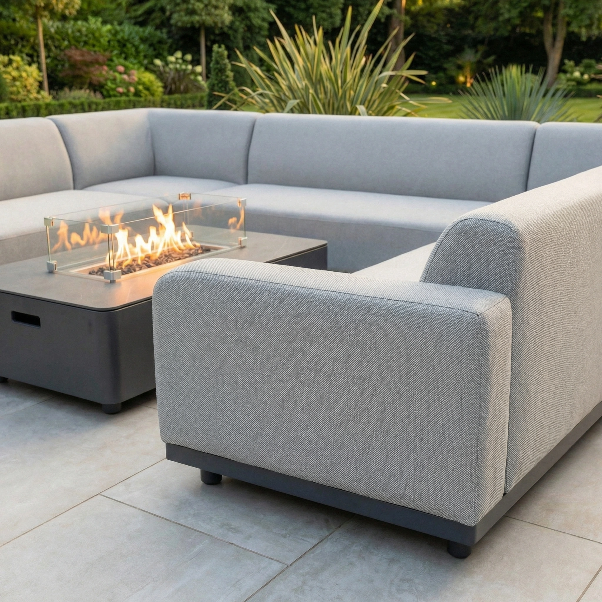 The Luna U-Shape Outdoor Fabric Sofa Set with Firepit Coffee Table in Oyster Grey is showcased on a tiled patio with a garden backdrop.