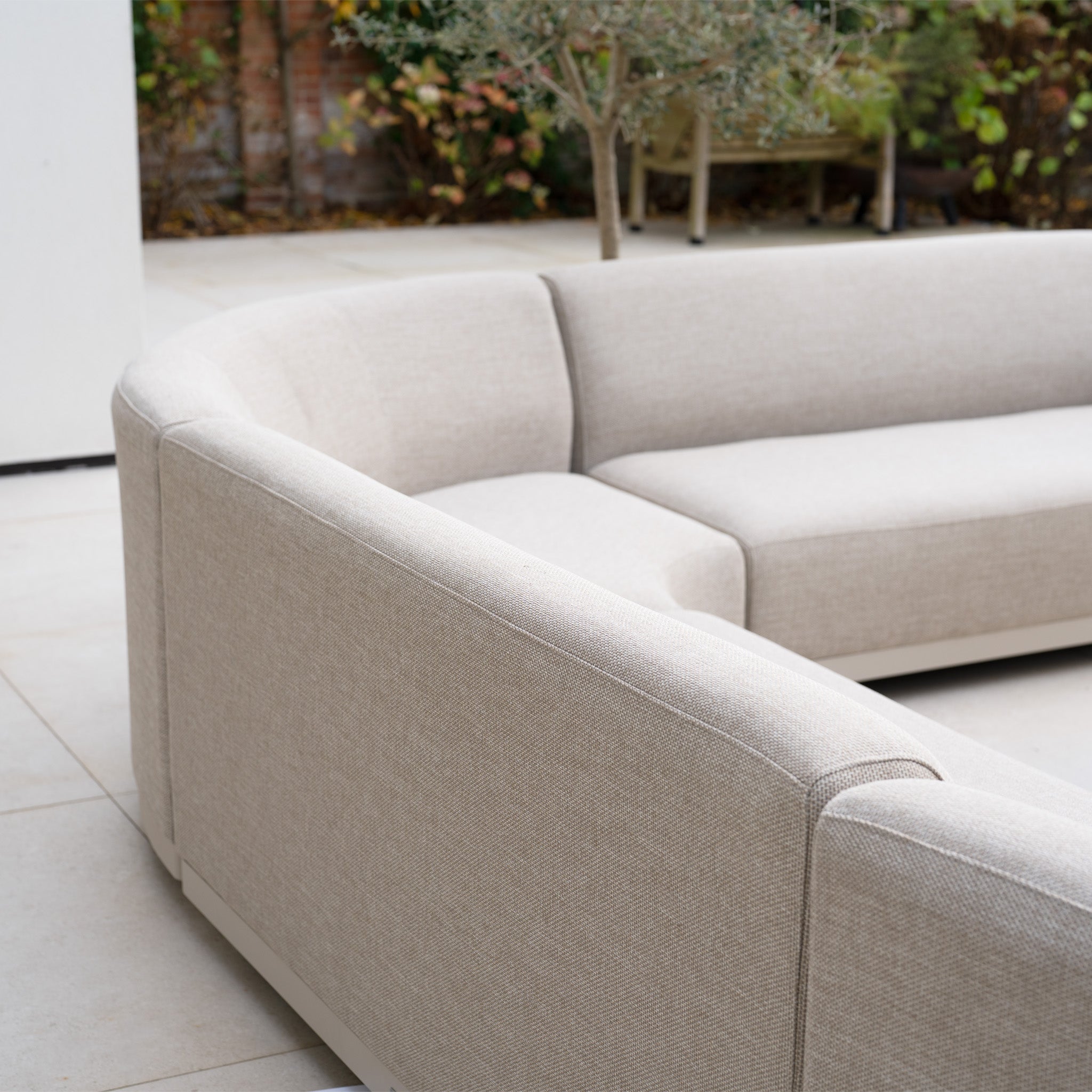 The Luna Outdoor Fabric Curved U-Shape Sofa Set with Altura Rising Table in Fawn sits on a tiled patio among lush plants—an elegant, all-weather garden feature ideal for stylish outdoor relaxation.