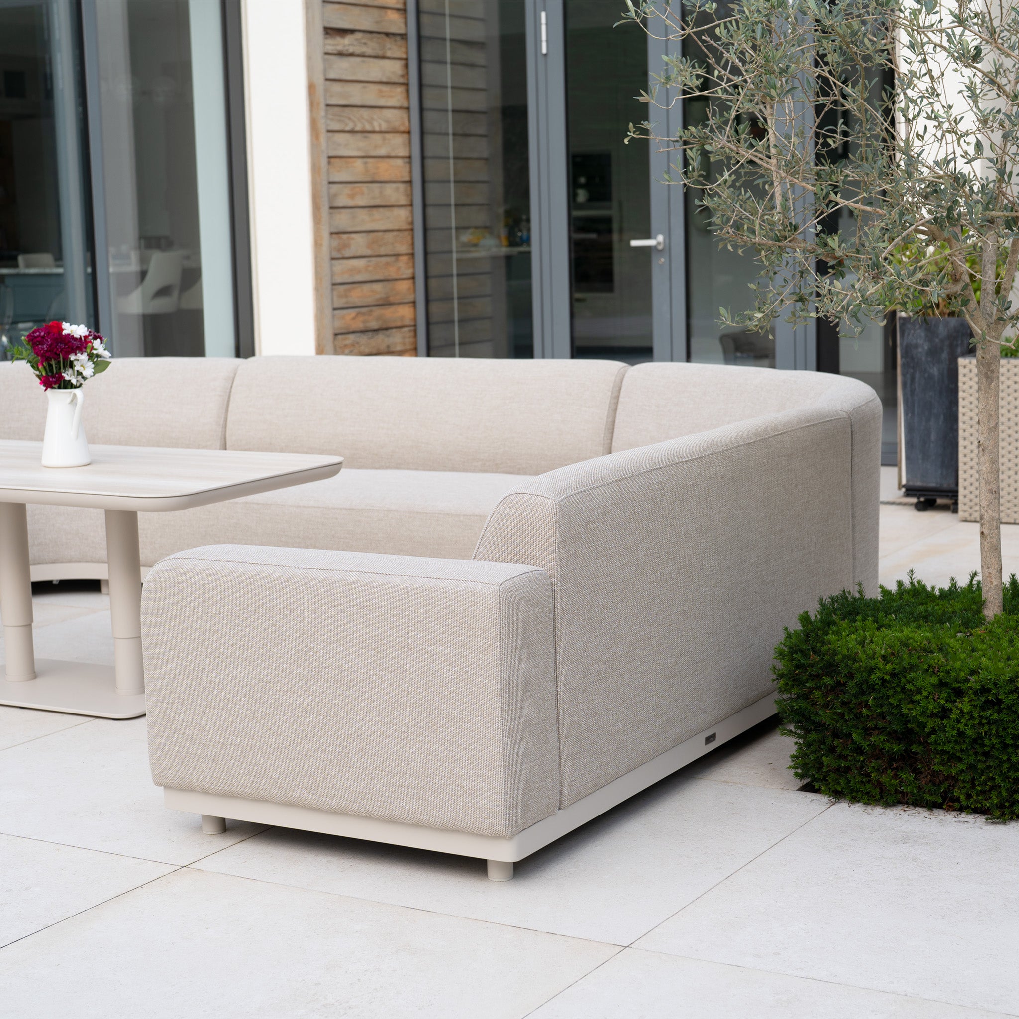 The Luna Outdoor Fabric Curved U-Shape Sofa Set with Altura Rising Table in Fawn offers perfect all-weather garden comfort—ideal for relaxing outdoors on your patio, next to green plants and glass doors.