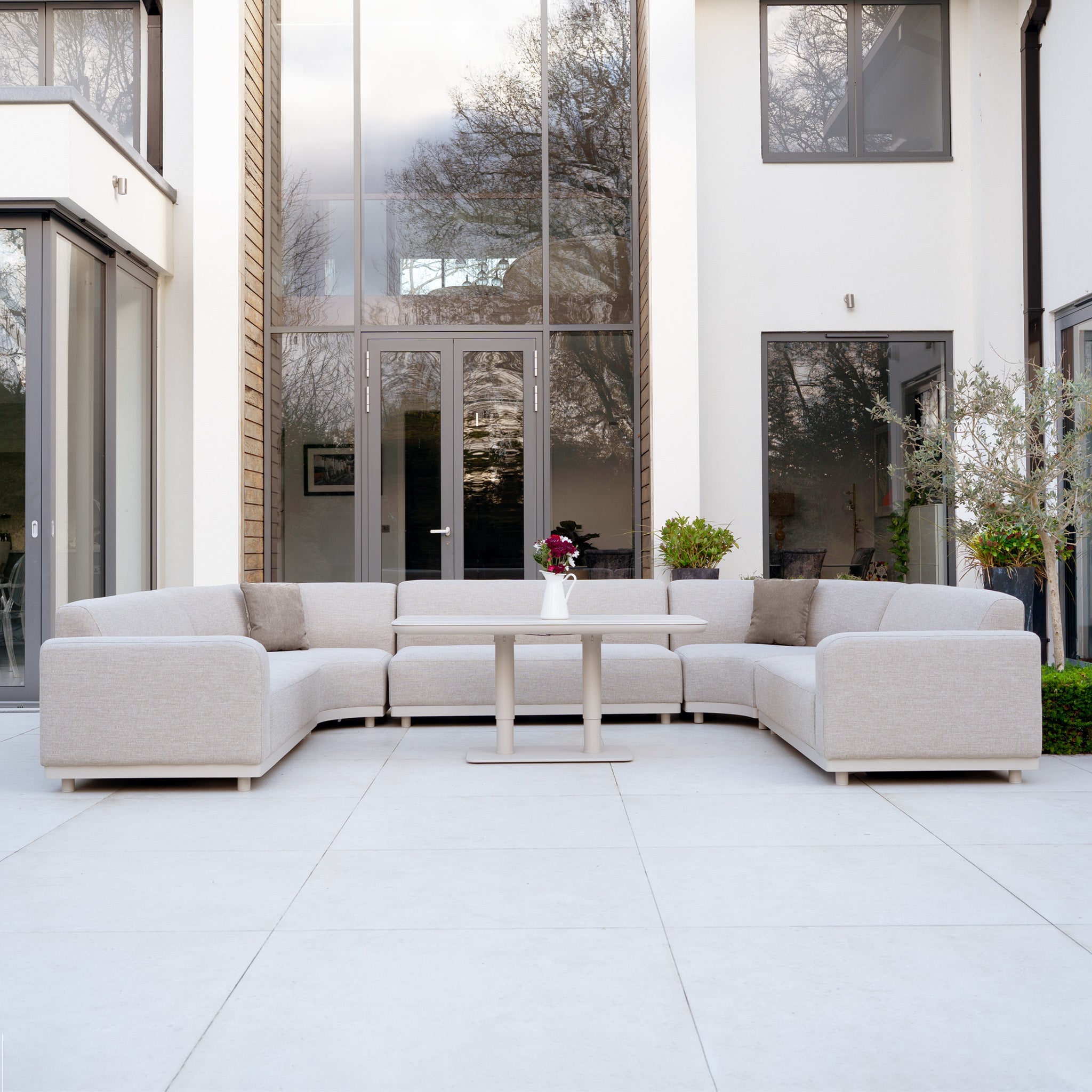 Modern patio featuring the Luna Outdoor Fabric Curved U-Shape Sofa Set in Fawn with Altura Rising Table amid plants, all-weather garden furniture set before large glass doors.