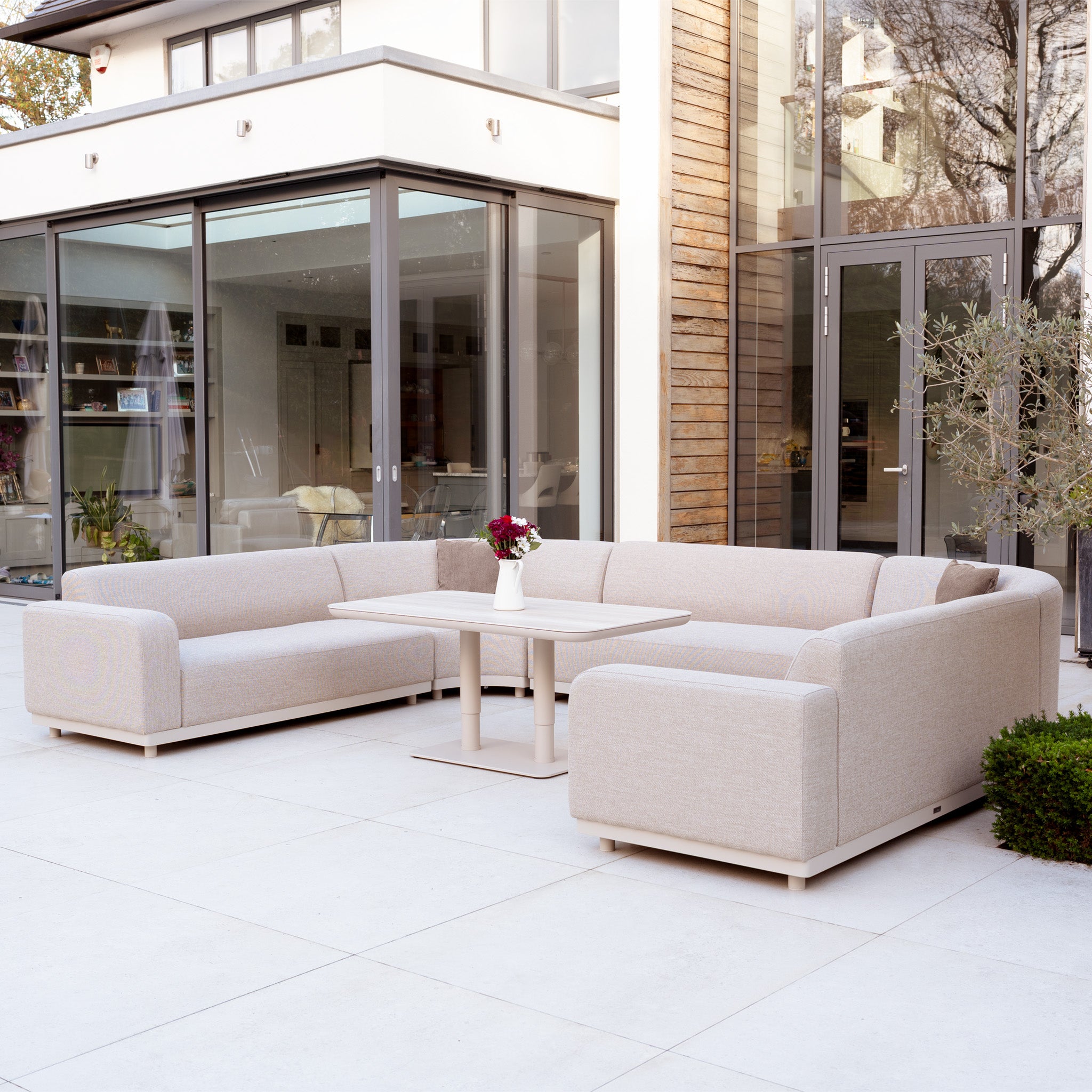 Modern patio with the Luna Outdoor Fabric Curved U-Shape Sofa Set and Altura Rising Table in Fawn beside a glass-walled house—a perfect showcase of stylish, all-weather garden furniture.