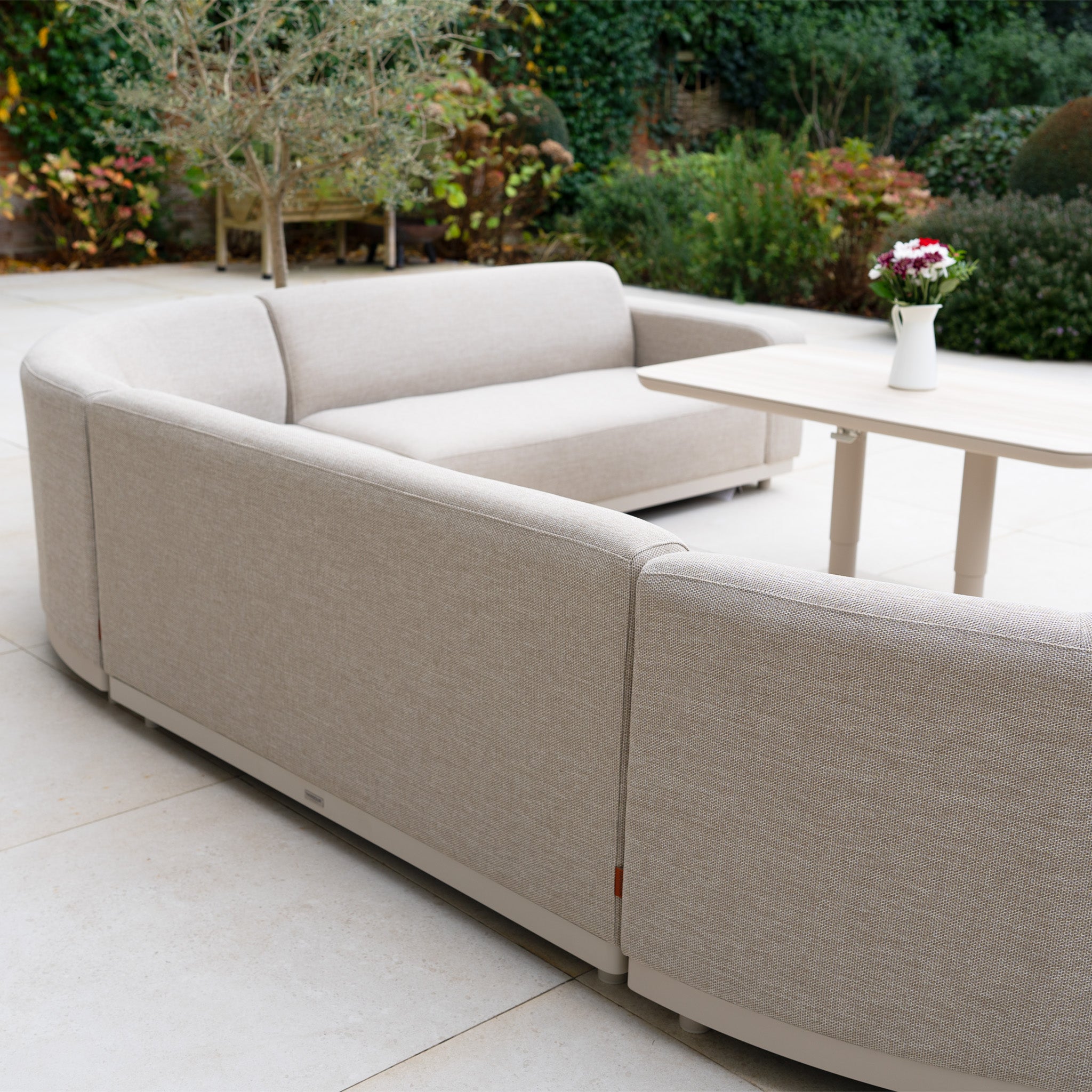 Curved Luna Outdoor Fabric U-Shape Sofa Set with Altura Rising Table in Fawn on a patio, adorned with a vase of flowers and greenery—an elegant, all-weather addition to your garden furniture collection.
