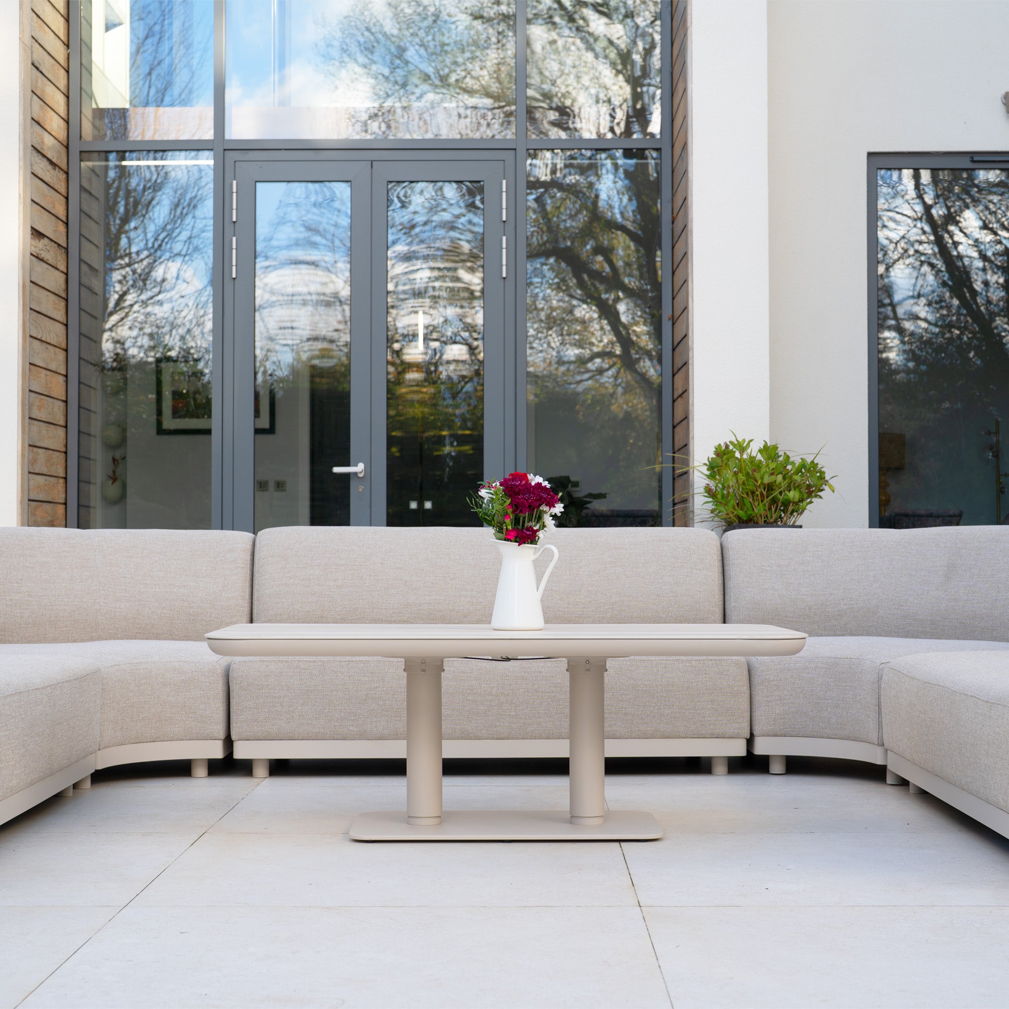Modern patio featuring the Luna Outdoor Fabric Curved U-Shape Sofa Set with Altura Rising Table in Fawn—stylish, all-weather garden furniture perfect for relaxing by large glass doors.