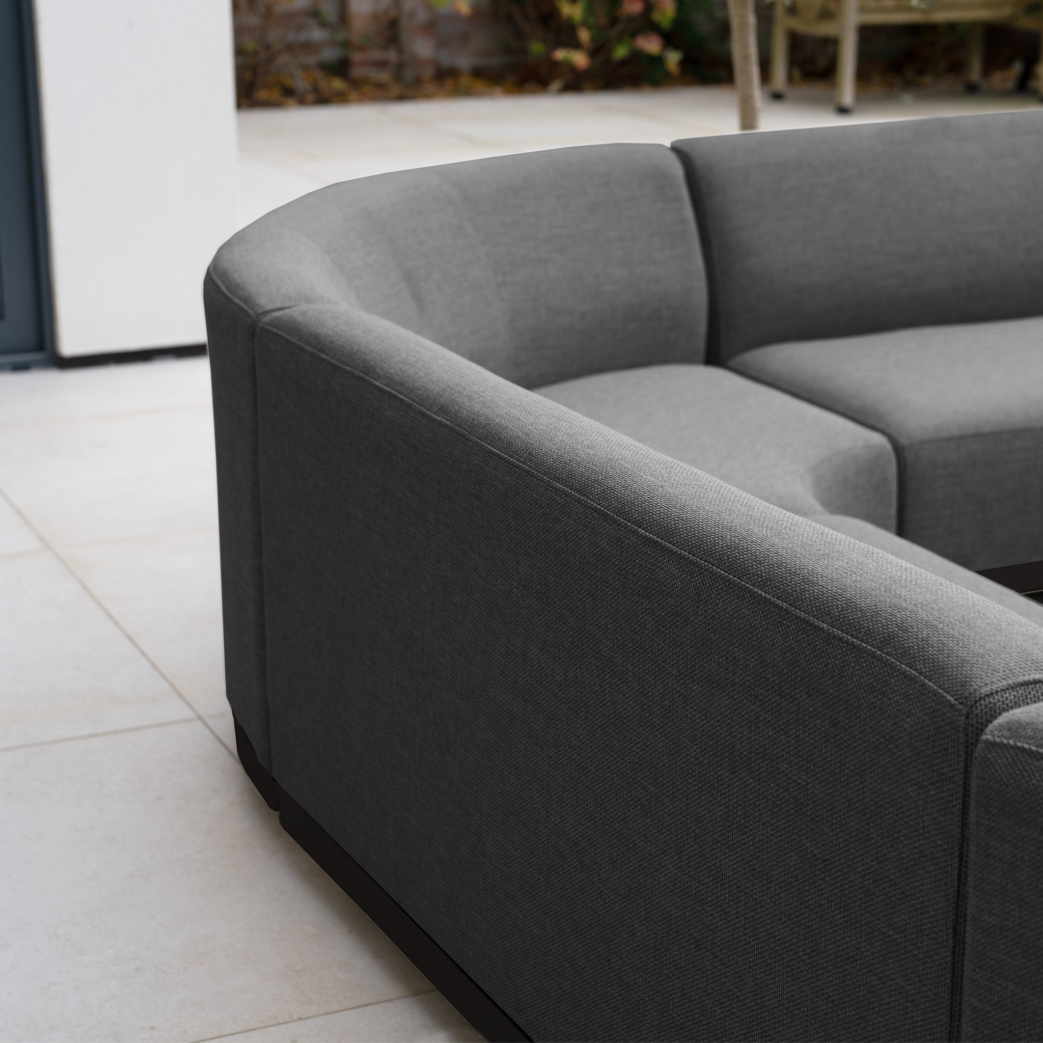 The Luna Outdoor Fabric Curved U-Shape Sofa Set with Altura Rising Table in Grey sits on light tiles, with all-weather garden furniture and outdoor plants in the background.