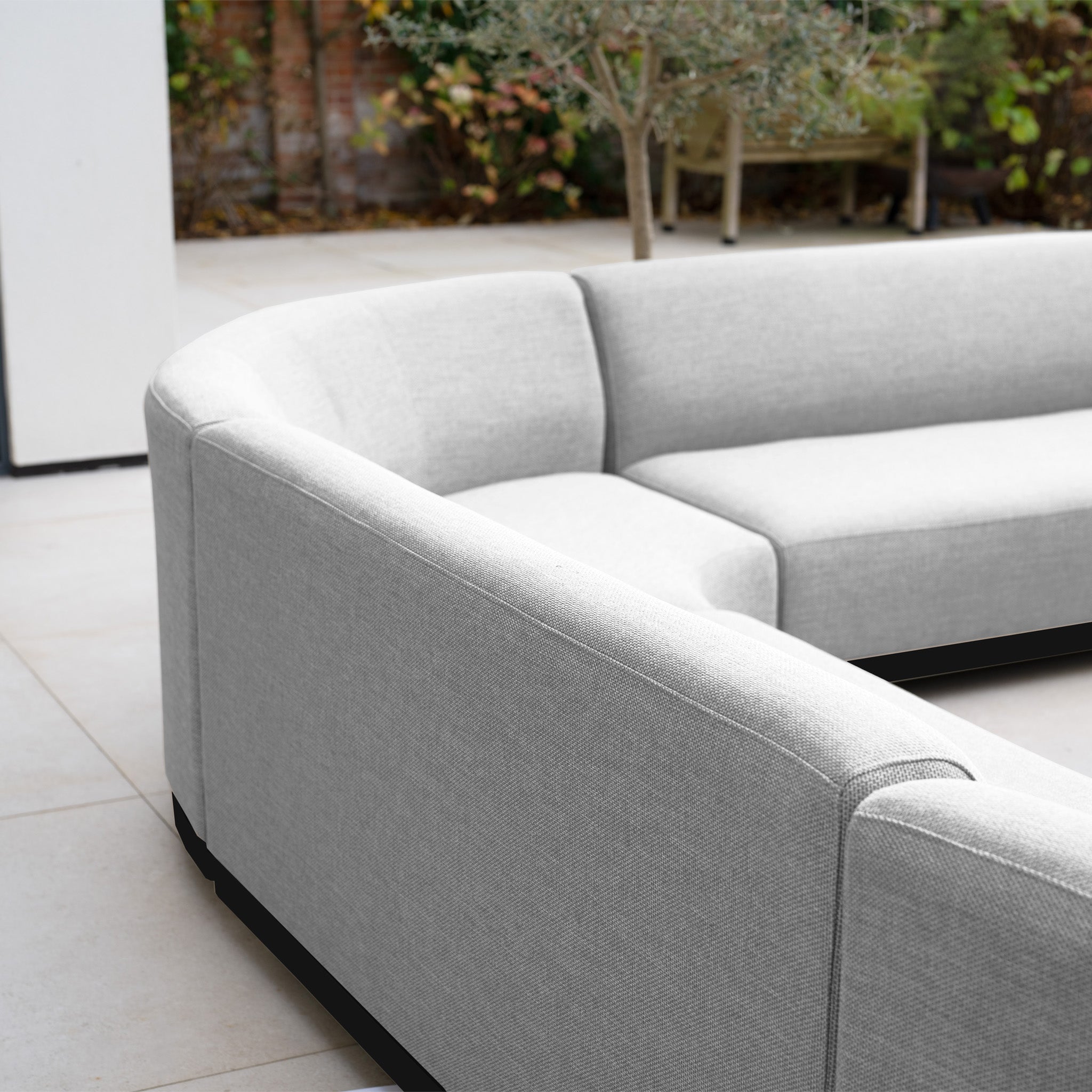 The Luna Outdoor Fabric Curved U-Shape Sofa Set with Altura Rising Table in Oyster Grey provides stylish, all-weather seating for any outdoor setting on your patio, surrounded by greenery and a tree.