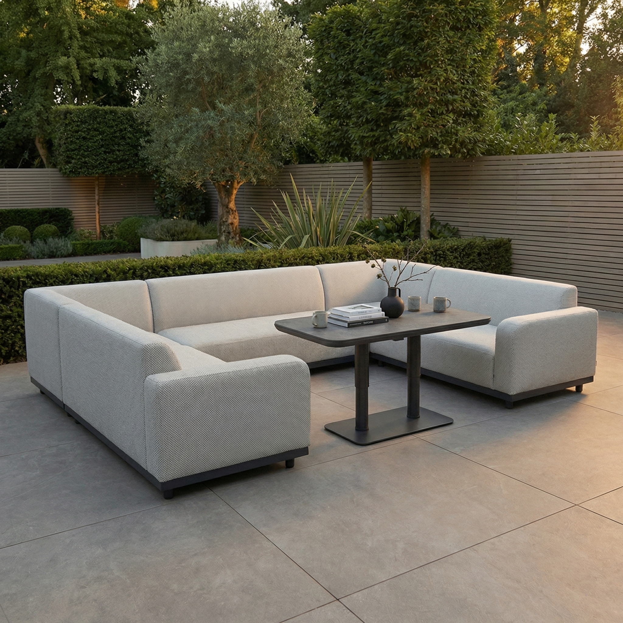 Modern outdoor patio featuring the Luna U-Shape Outdoor Fabric Sofa Set with Altura Rising Table in Oyster Grey, crafted with rain-resistant fabric and set on a tiled floor surrounded by lush greenery.