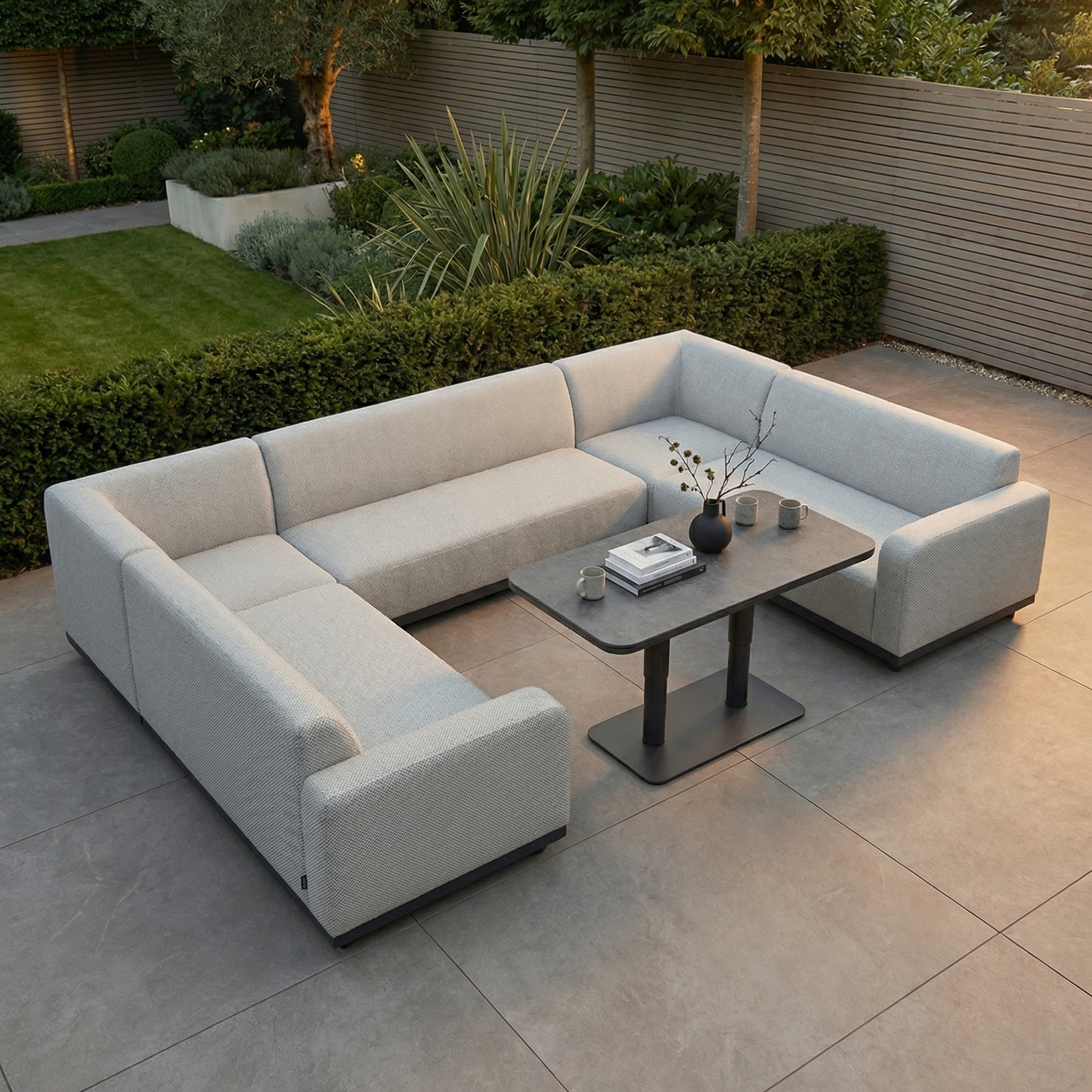 The Luna U-Shape Outdoor Fabric Sofa Set in Oyster Grey, featuring an Altura Rising Table, sits on a stone patio and is surrounded by lush greenery. Its rain-resistant fabric makes it ideal for outdoor garden use.