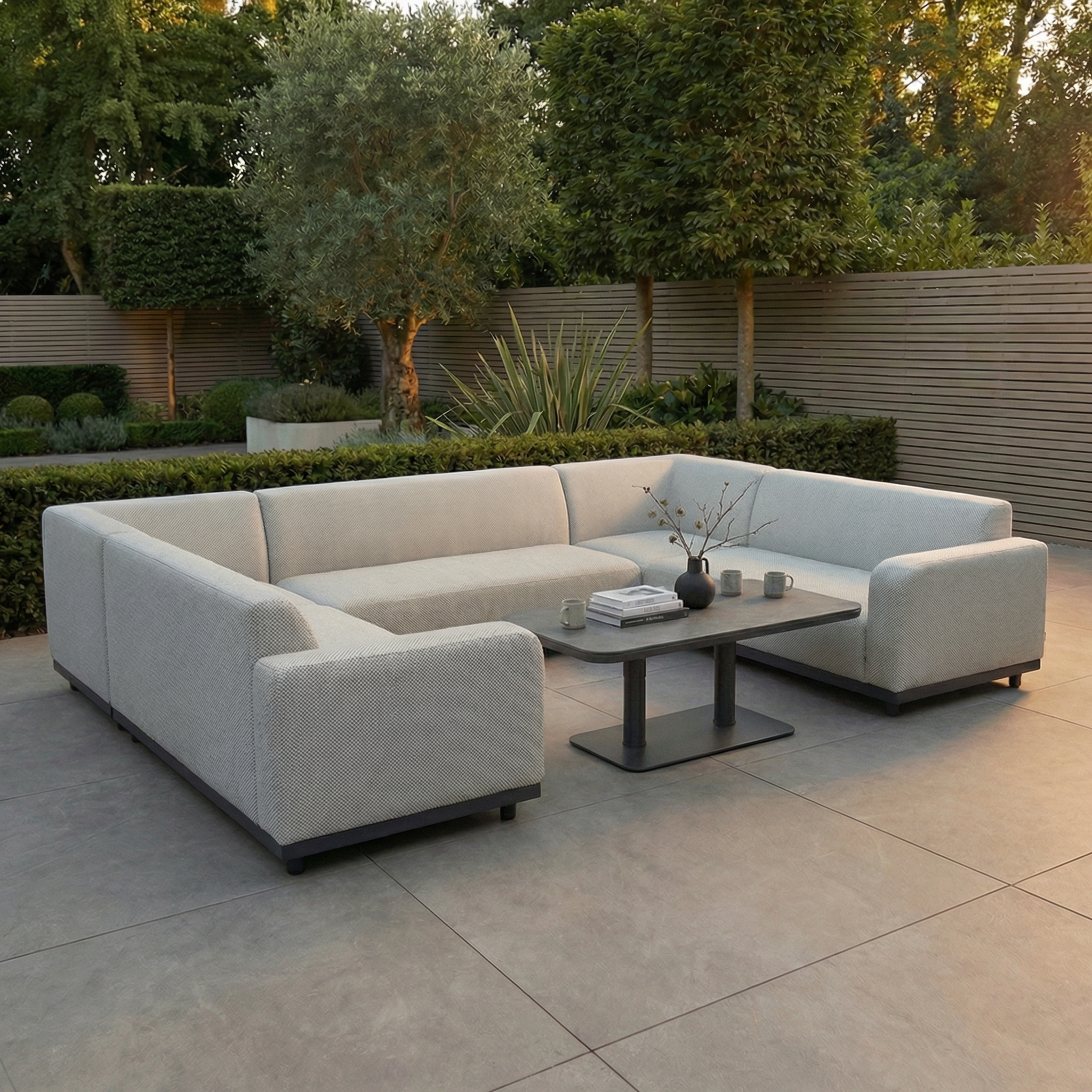Modern outdoor patio with the Luna U-Shape Outdoor Fabric Sofa Set in Oyster Grey and Altura Rising Table, arranged on large stone tiles and surrounded by lush greenery.