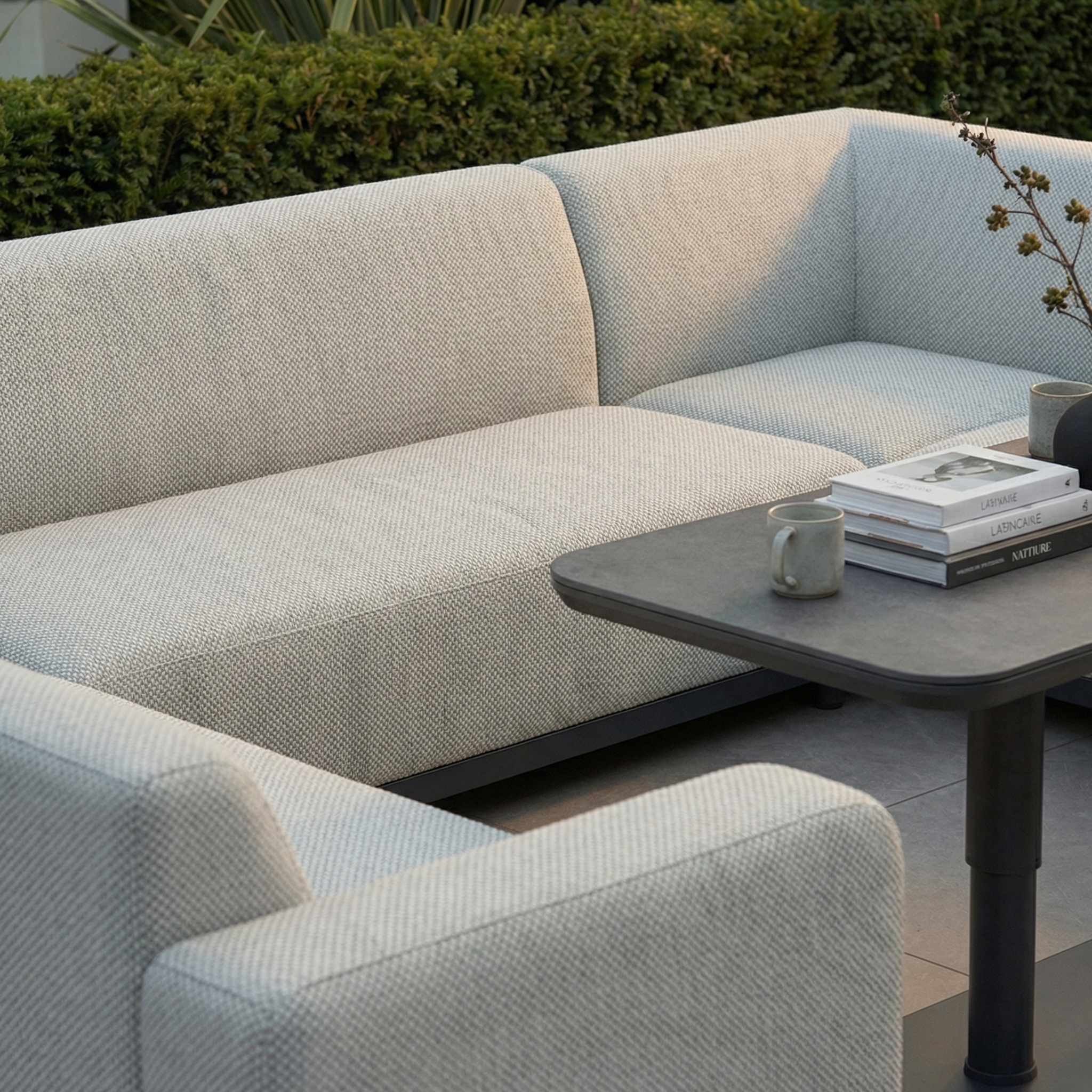 Create a modern outdoor retreat with the Luna U-Shape Outdoor Fabric Sofa Set and Altura Rising Table in Oyster Grey—rain-resistant, stylish garden furniture perfect for relaxing among lush greenery with books and cups.