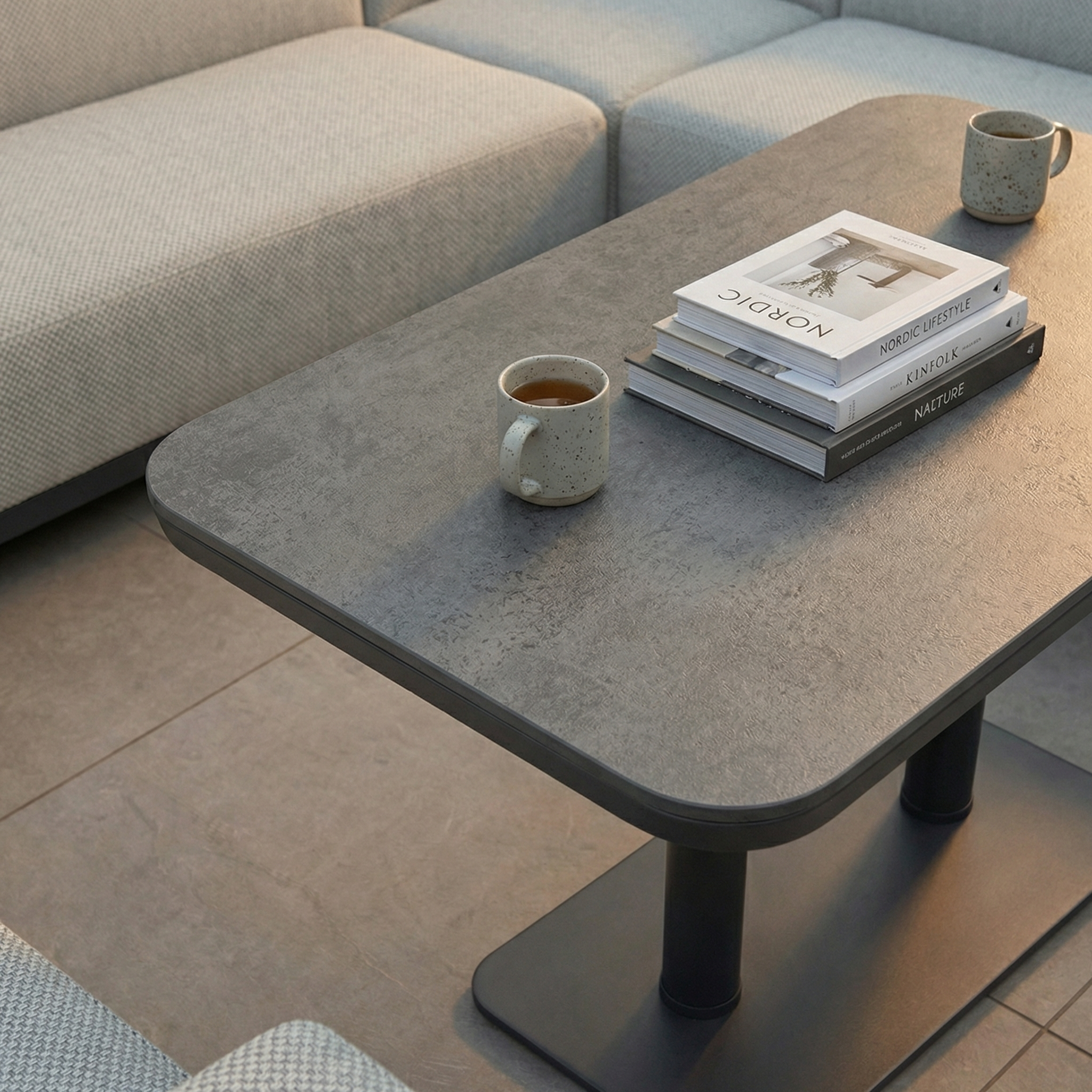 The Luna U-Shape Outdoor Fabric Sofa Set in Oyster Grey features rain-resistant sofas and an Altura rising coffee table, shown with books and two mugs in a cozy living room setting.
