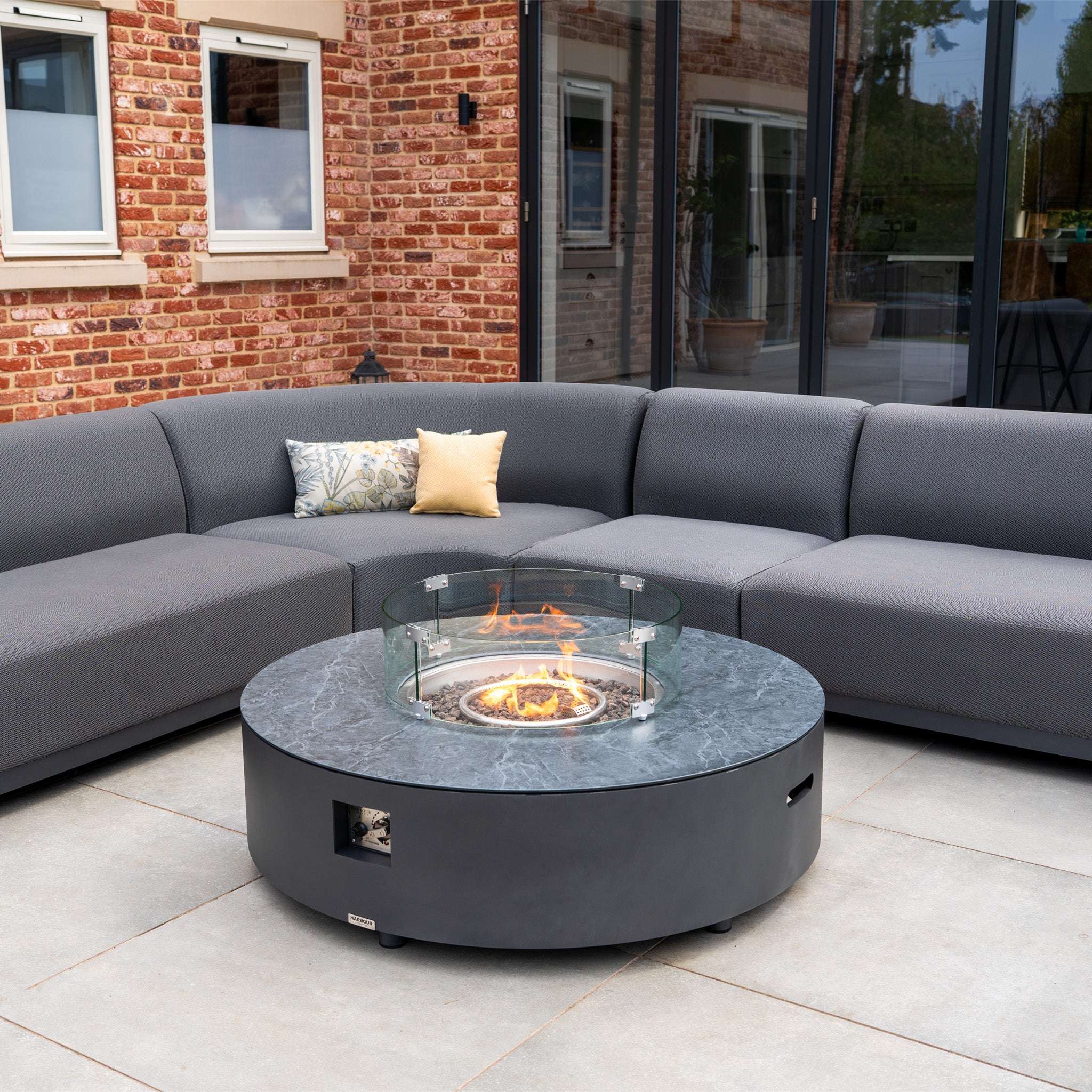 Modern patio with the Luna U-Shape Outdoor Fabric Curved Corner Dining set in grey, featuring a curved sofa and round firepit coffee table on stone tile flooring against brick walls.