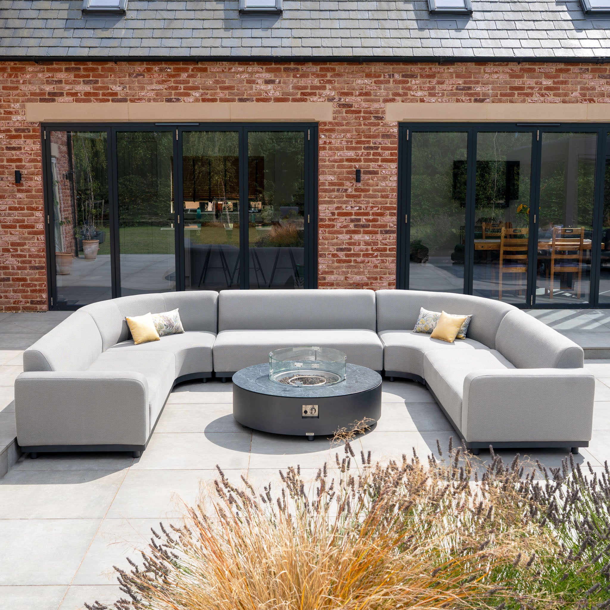 The Luna U-Shape Outdoor Fabric Curved Corner Group with Round Firepit Coffee Table in Oyster Grey provides stylish patio seating, complete with throw pillows and a cozy firepit table by the glass doors.