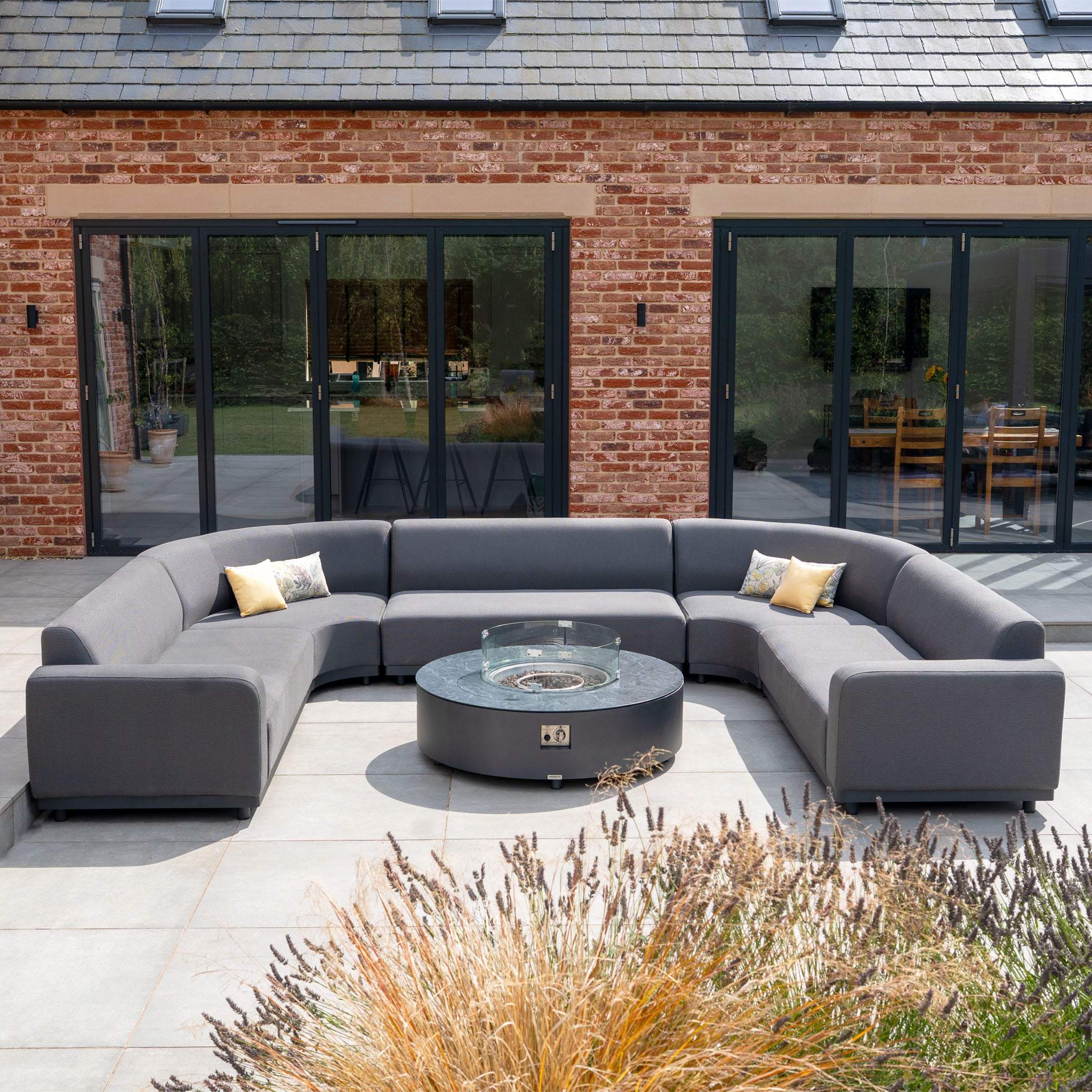 The Luna U-Shape Outdoor Fabric Curved Corner Dining Set in Grey features a spacious sofa and a round firepit coffee table, perfect for stylish patio gatherings in front of glass doors and a classic brick house.