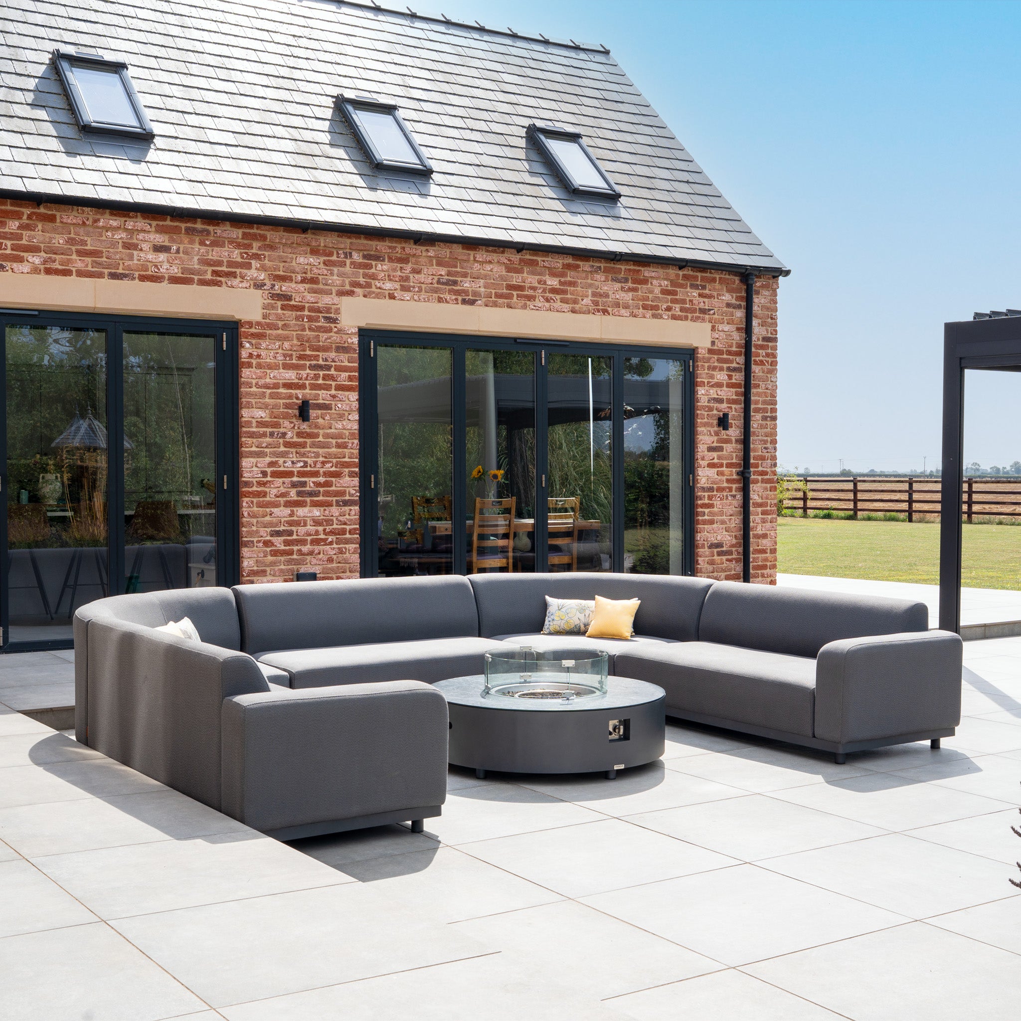 The Luna U-Shape Outdoor Fabric Curved Corner Dining Set with Round Firepit Coffee Table in Grey creates a stylish, inviting space for relaxing or dining outdoors by your brick house on a sunny patio.