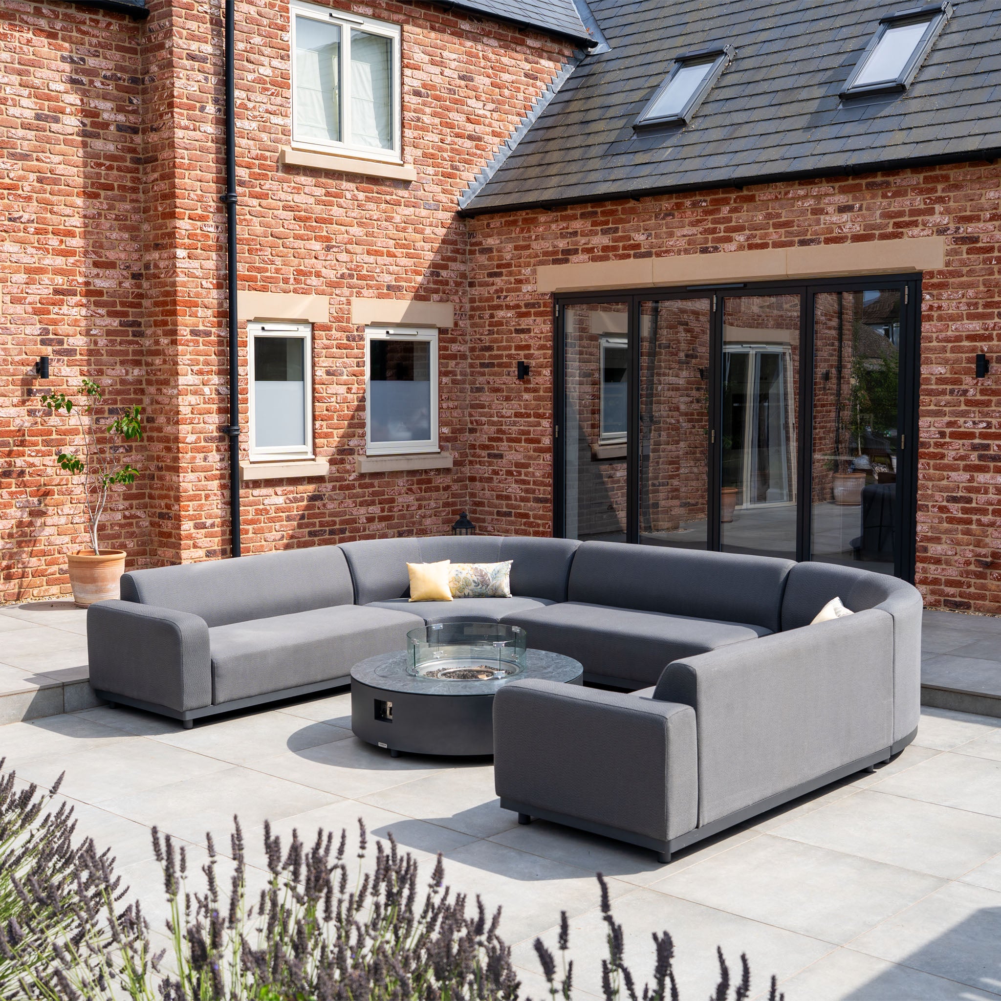 The Luna U-Shape Outdoor Fabric Curved Corner Dining with Round Firepit Coffee Table in Grey creates a cozy, luxurious patio setup beside a red brick house with glass sliding doors.