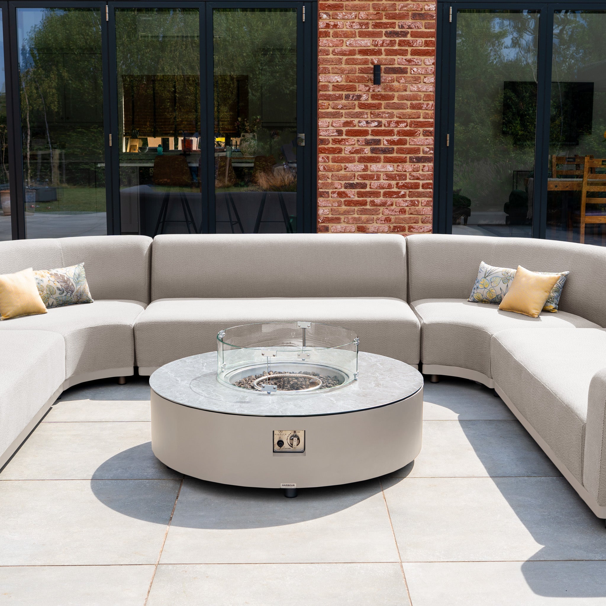 The Luna U-Shape Outdoor Fabric Curved Corner Group in Fawn with plush cushions and a round firepit coffee table is arranged on a patio in front of glass doors.