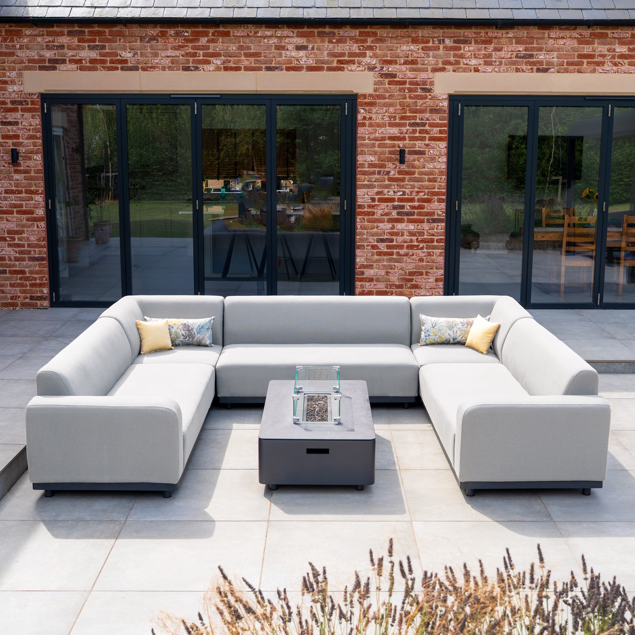The Luna U-Shape Outdoor Fabric Sofa Set with Firepit Coffee Table in Oyster Grey is arranged on a patio next to a brick house.