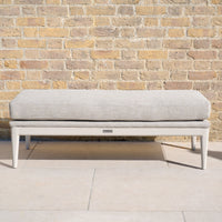 The Luna Bench in Fawn, a beige upholstered all-weather bench with white legs, adds elegant outdoor seating against a tan brick wall on a stone patio.
