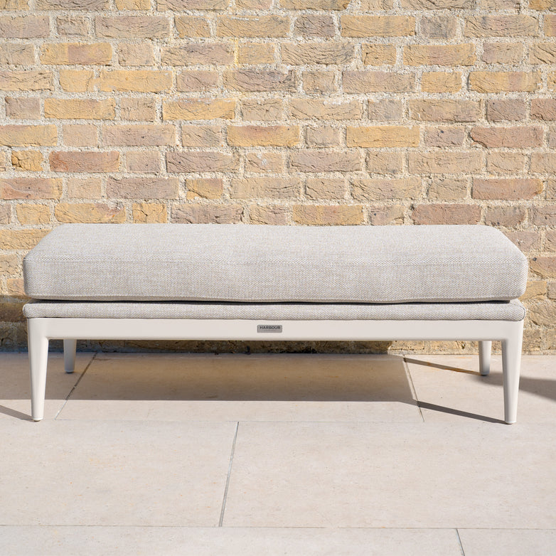 The Luna Bench in Fawn, a beige upholstered all-weather bench with white legs, adds elegant outdoor seating against a tan brick wall on a stone patio.