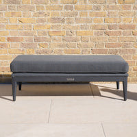 The Luna Bench in Grey, featuring a modern design and textured cushion, is placed against a light brick wall on a tiled patio.