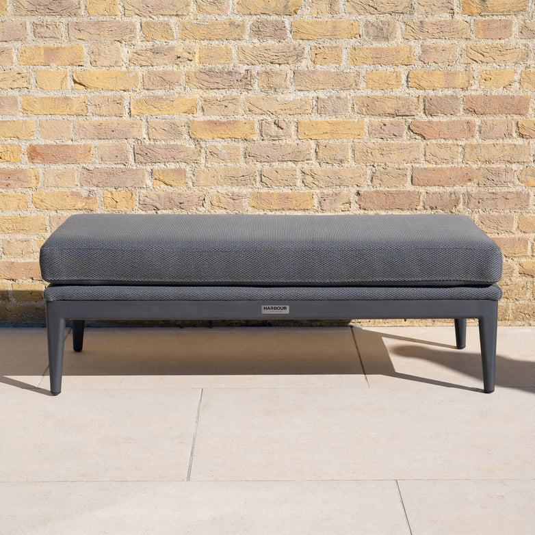 The Luna Bench in Grey, featuring a modern design and textured cushion, is placed against a light brick wall on a tiled patio.